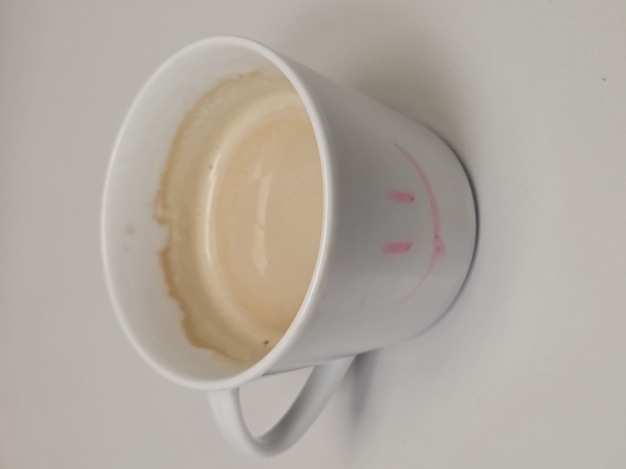 A top-down angled close-up shot shows a partially consumed beverage in a white mug resting on a white surface. The mug has a simple handle and a pink, childlike drawing of a smile on its side. Coffee stains are visible inside the mug, near the rim. The remaining liquid appears light brown, likely coffee with milk. The background is a plain white surface. The image provides insufficient information to determine the location in Varna, Bulgaria. There are no people or interactions visible.