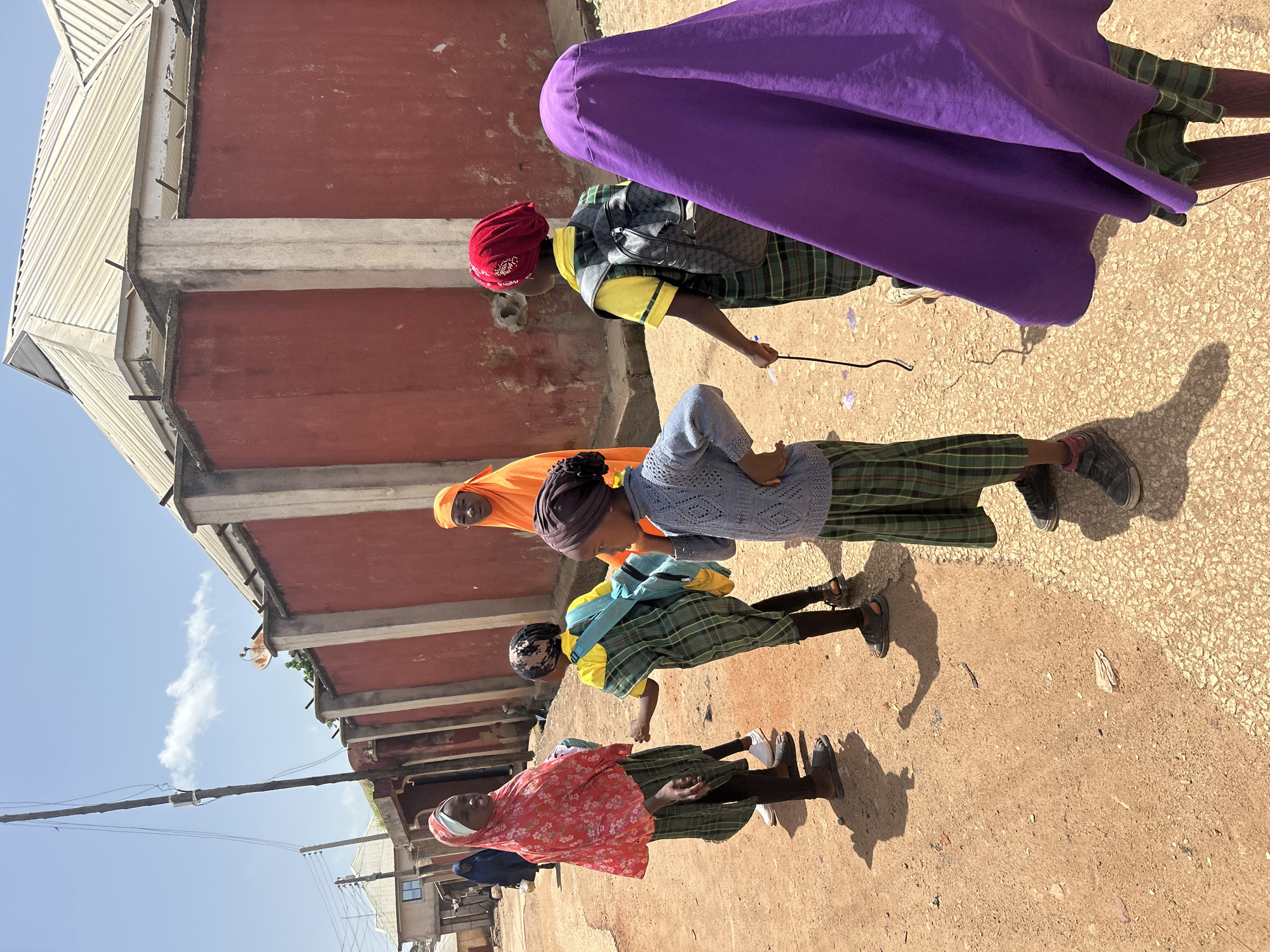 In Jos, Nigeria, several young girls, likely students, are gathered on a dirt road in front of a large, red-walled building with a corrugated metal roof. The building appears to be a residential or commercial property. One girl in the foreground wears a red headscarf with a floral pattern, a green plaid skirt, and black leggings. Next to her, a girl in a yellow top and green plaid skirt has a blue backpack. Another girl in the foreground is seen from the side, wearing a blue knitted sweater, a green plaid skirt, and a dark headscarf. In the background, a woman in an orange hijab stands with another girl in a purple covering. The environment is characterized by a dry, dusty ground and a bright, sunny sky. The overall scene suggests a moment of interaction or waiting, possibly before or after school.