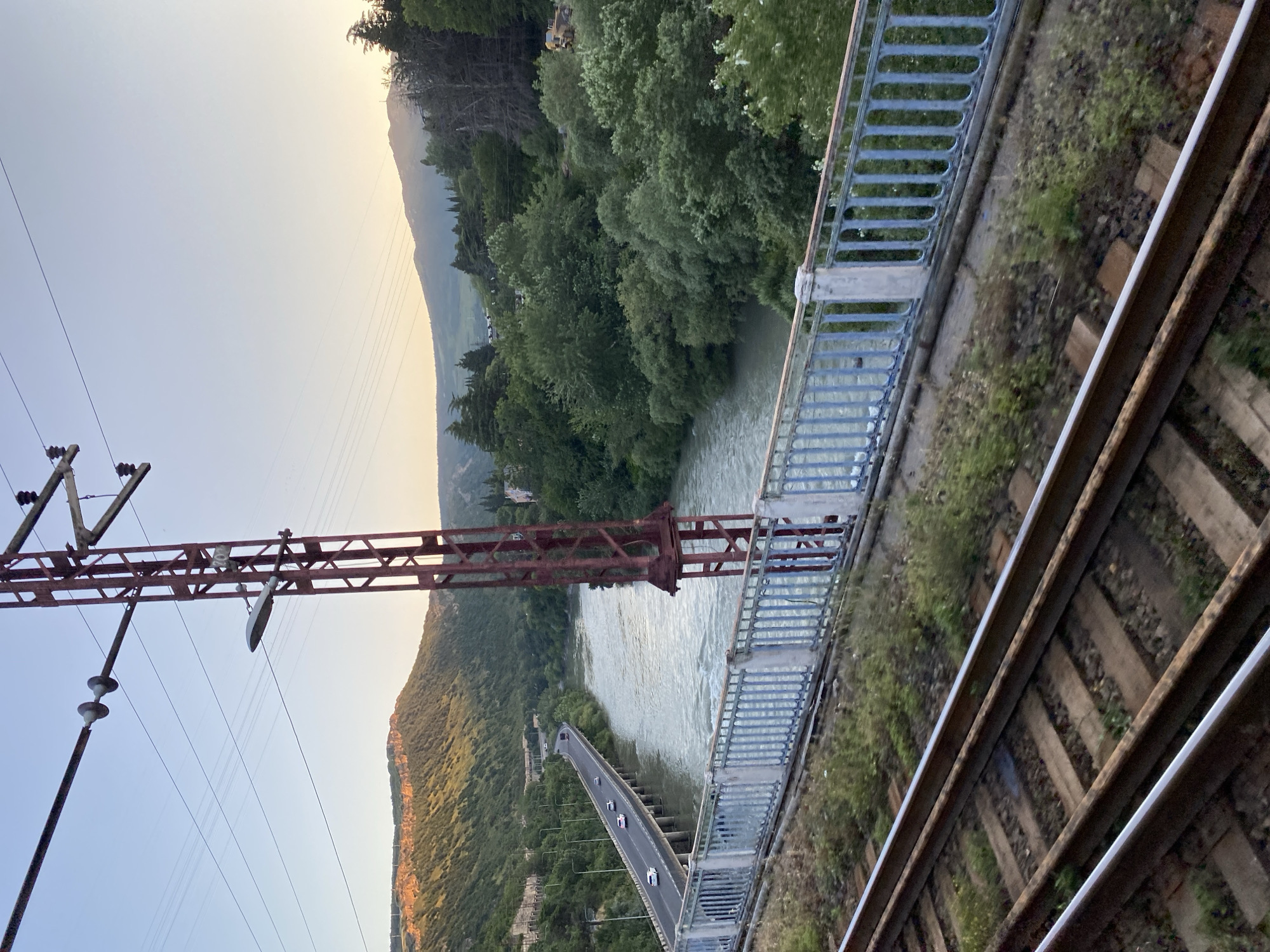 The image shows a view from a bridge in Tbilisi, Georgia, looking across a river and a valley. The scene is dominated by lush green trees on the hillsides and a wide, fast-flowing river. In the foreground, there are railroad tracks, suggesting a railway bridge or line runs parallel to the one depicted. Adjacent to the railroad tracks is another bridge with a metal railing, likely a pedestrian or road bridge. Further in the distance, a longer, elevated road bridge spans the river, with several cars visible on it.
A significant element of the scene is a large, rusty red metal structure that appears to be a pylon or support for overhead power lines, as several wires are suspended from it. This structure is positioned between the viewer and the river. The sky is a pale, hazy blue, transitioning to a warmer, golden hue near the horizon, indicating it is either early morning or late afternoon. The sun is not directly visible, but its light casts a soft glow on the landscape, highlighting the mountains in the distance. The overall mood is peaceful and scenic, with a sense of natural beauty and infrastructure coexisting. There is no visible text in the image.