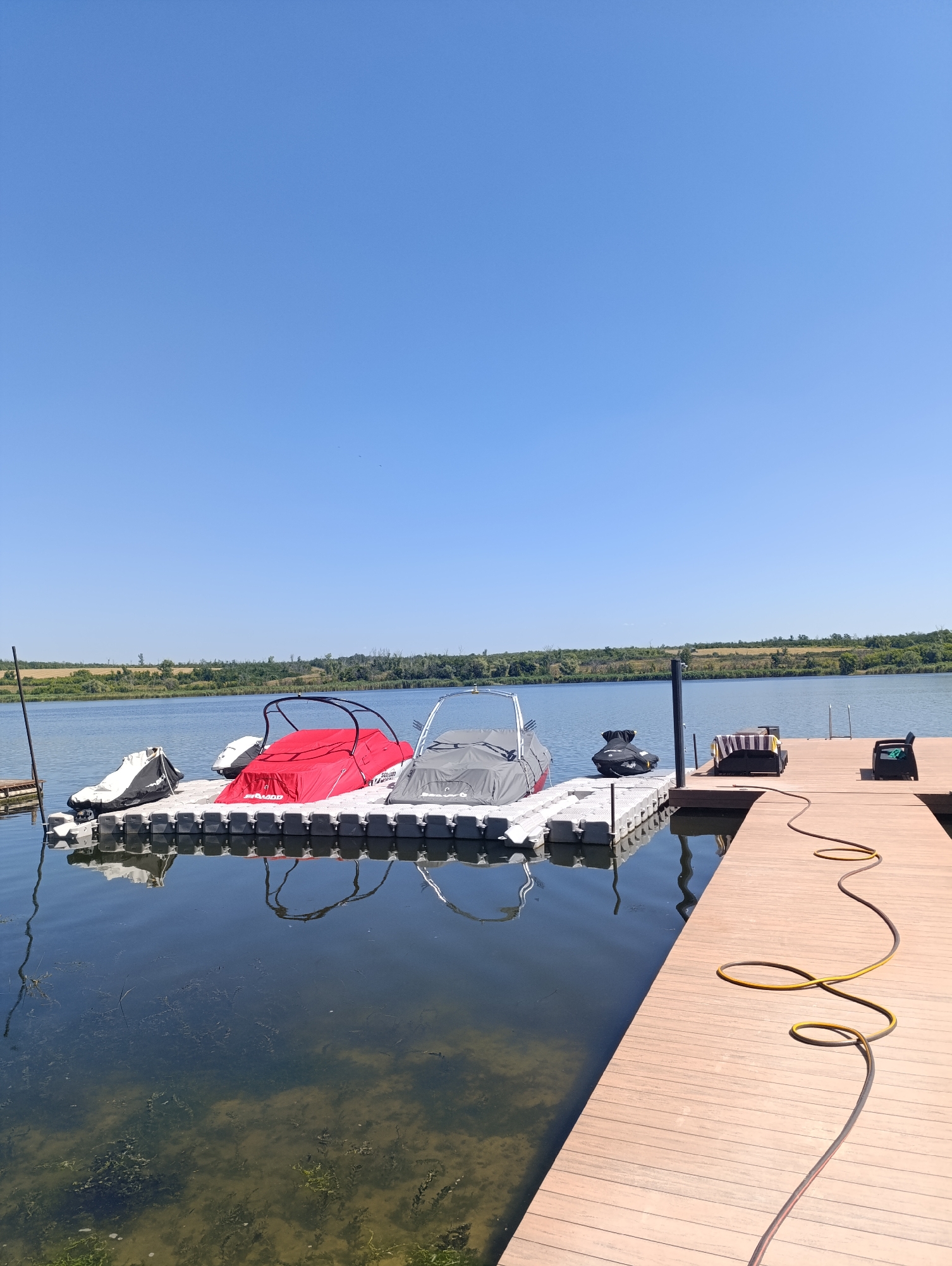 The image shows a wooden dock extending into a calm body of water. The dock is made of wooden planks and has a yellow hose coiled on its surface. Adjacent to the main dock is a floating modular dock system. On this floating dock, there are several watercraft, some covered with tarps. One covered watercraft is predominantly red, and another is grey. Two jet skis are also present on the floating dock, one black and one white. The water appears clear with visible aquatic vegetation near the edge of the floating dock. In the background, a treeline borders the water under a clear blue sky. The scene is set in Bulatselivka, Ukraine.