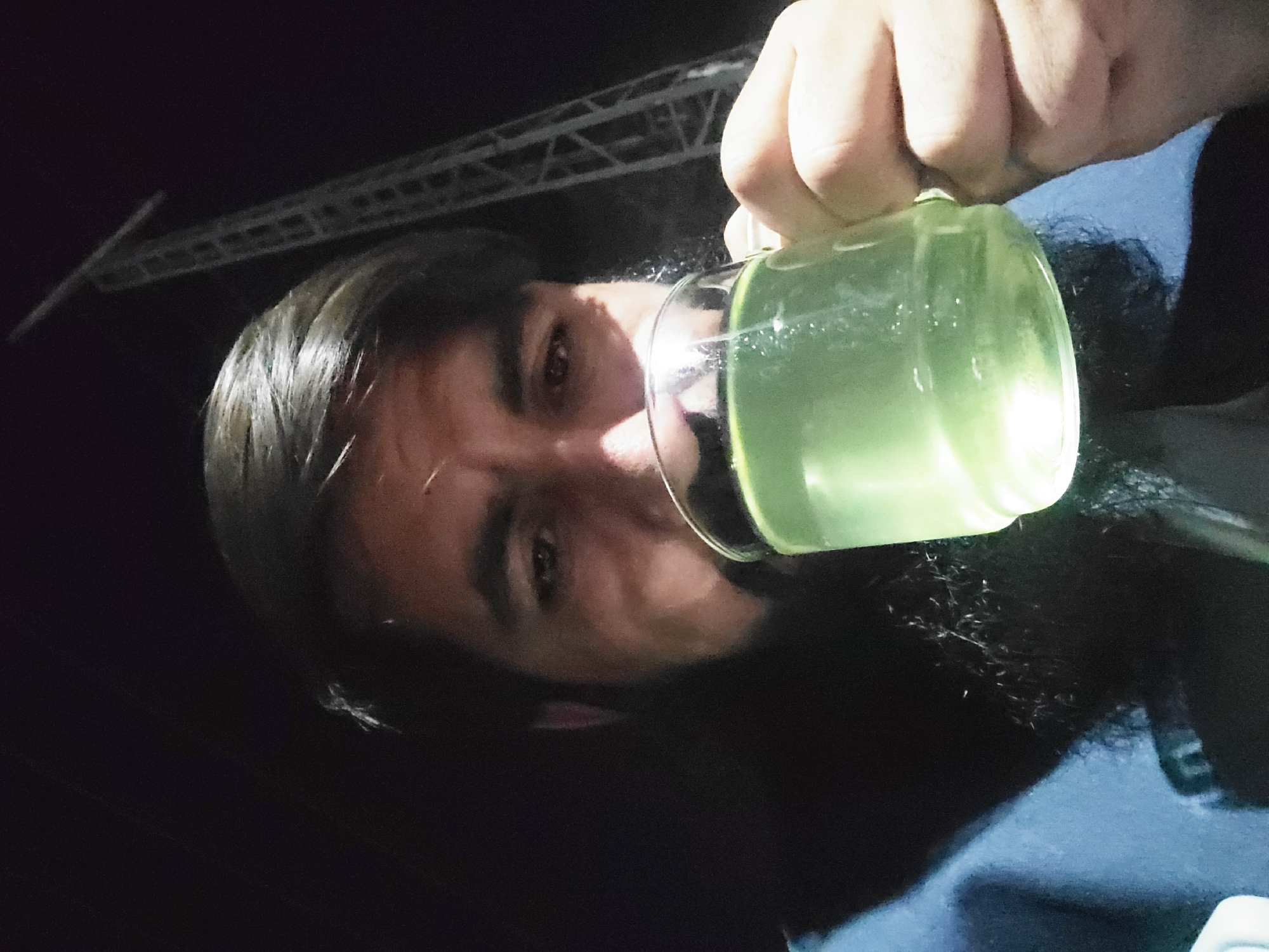 The image shows a close-up, low-angle shot of a person drinking from a glass, set against a dark background, likely at night in Quetta City Tehsil, Pakistan. The primary subject is a person, appearing male, with dark hair and visible facial features including an eye and eyebrow. Their right hand firmly grips a clear glass filled with a luminous, light green liquid, which they are actively consuming. The glass is positioned close to their mouth, obscuring part of their face.
The background is predominantly dark, indicating either nighttime or a dimly lit indoor environment. In the upper left portion of the image, a faint, blurred metallic structure, possibly scaffolding or a beam, is visible, offering a subtle clue about the surroundings. The liquid in the glass glows brightly, illuminating the glass itself and casting a soft, greenish light onto the person's face, which is a notable detail. No strong emotions are clearly discernible from the person's expression, but they appear focused on drinking. There is no visible text within the image.