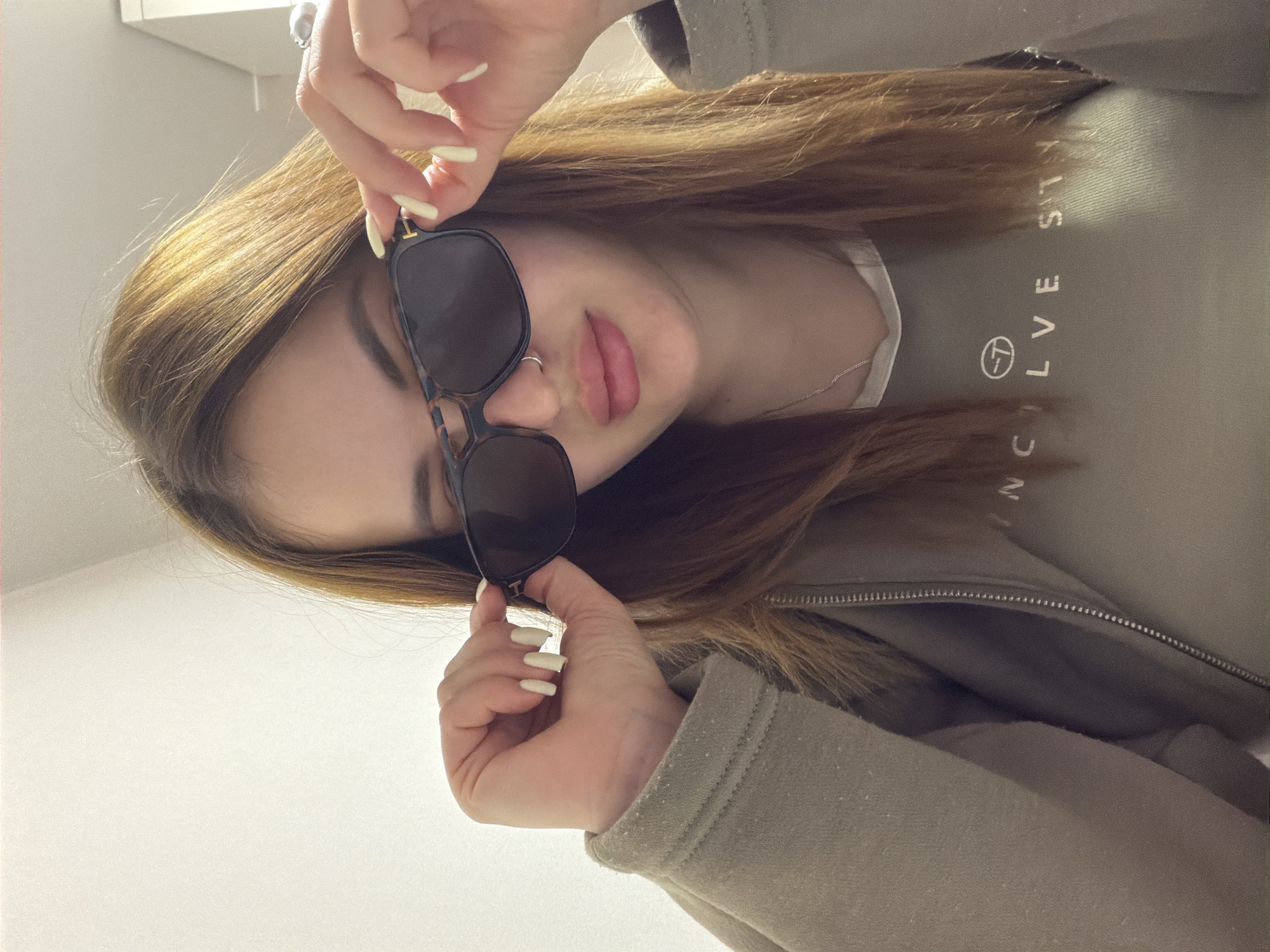 A woman with light brown hair and long, light-colored nails is holding dark sunglasses. She is wearing a light brown sweatshirt with "VOLVE SITY" printed on it. The background is a plain white wall. The location is not identifiable as Krasnodar, Russia from the provided image.