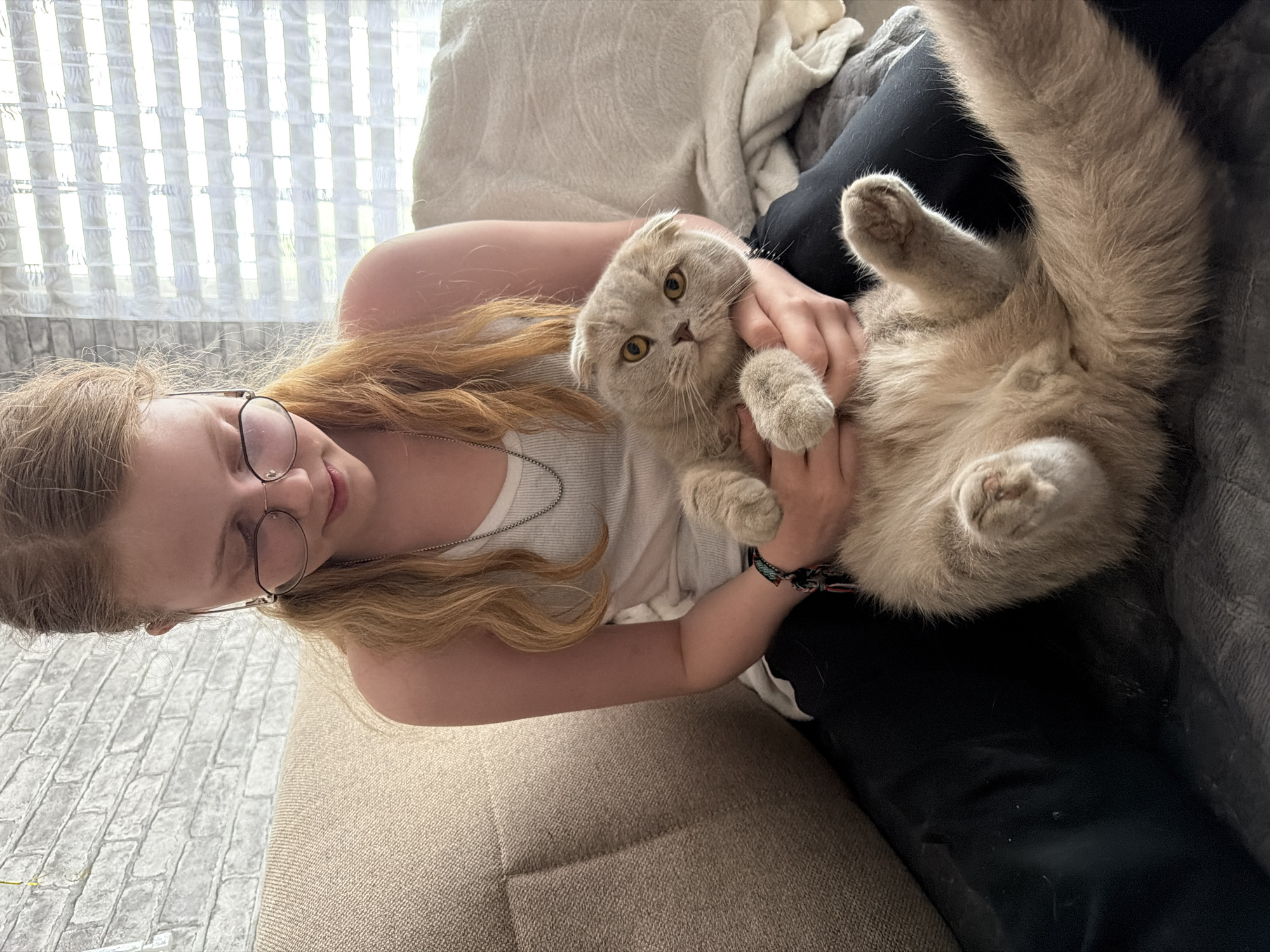 A fair-skinned, blonde-haired person wearing glasses is holding a light brown Scottish Fold cat belly-up. The person is wearing a white tank top and has a silver necklace. They are seated on a couch with a light brown cushion. Behind them is a window with a textured glass covering. A gray blanket or cushion is also visible. The location is Chaikovsky, Russia.