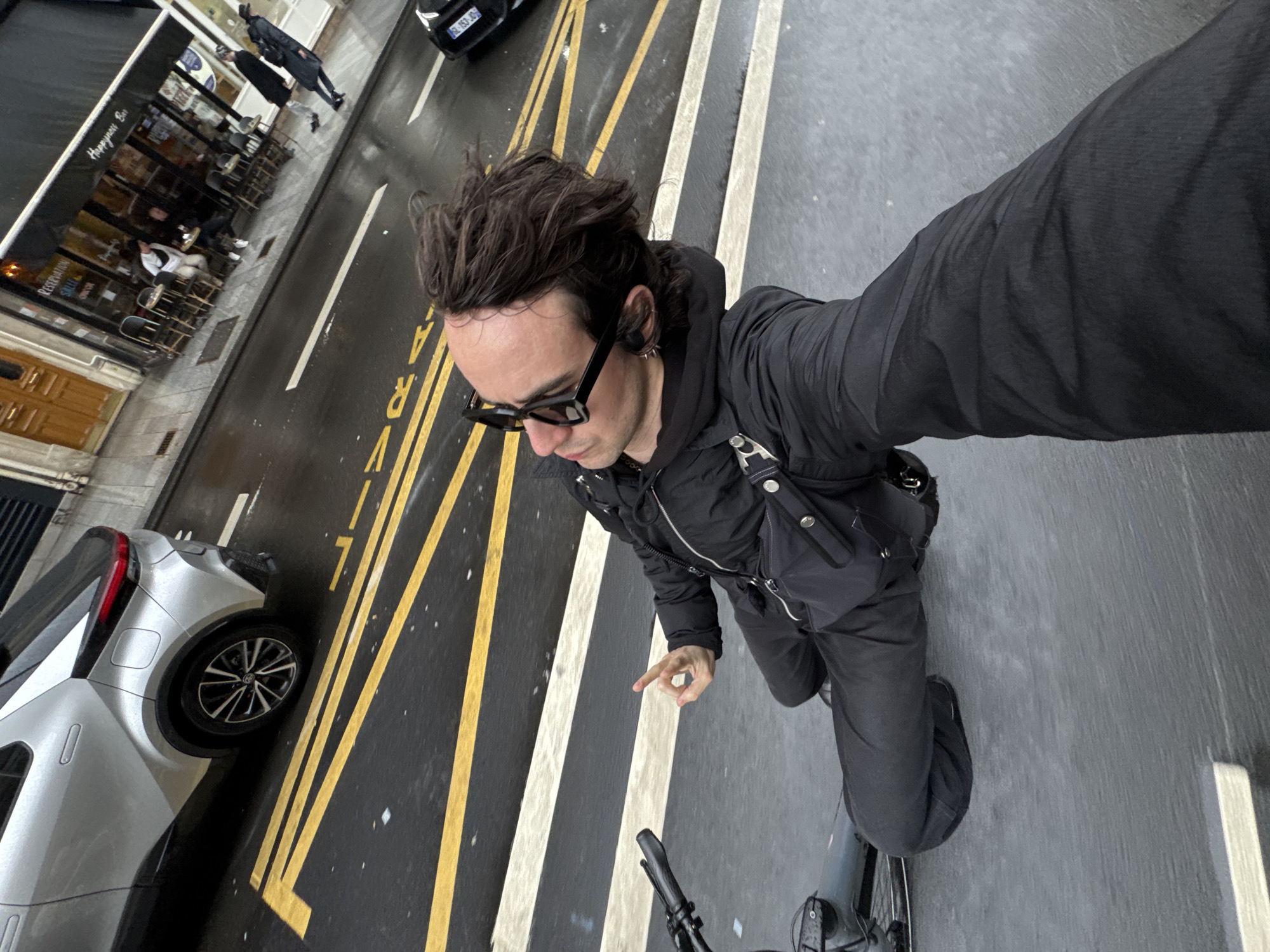 A man is riding a bicycle on a street in Paris, France. The street has yellow and white lines painted on it, indicating it is a designated lane. He is wearing a black jacket, black pants, and black sunglasses, and he has dark hair. He appears to be taking a selfie. To the left of the man is a silver car parked along the road. On the opposite side of the street, there is a restaurant or bar with outdoor seating. People are sitting at the tables. The street appears to be wet, suggesting it has recently rained. The buildings along the street are typical Parisian architecture.
