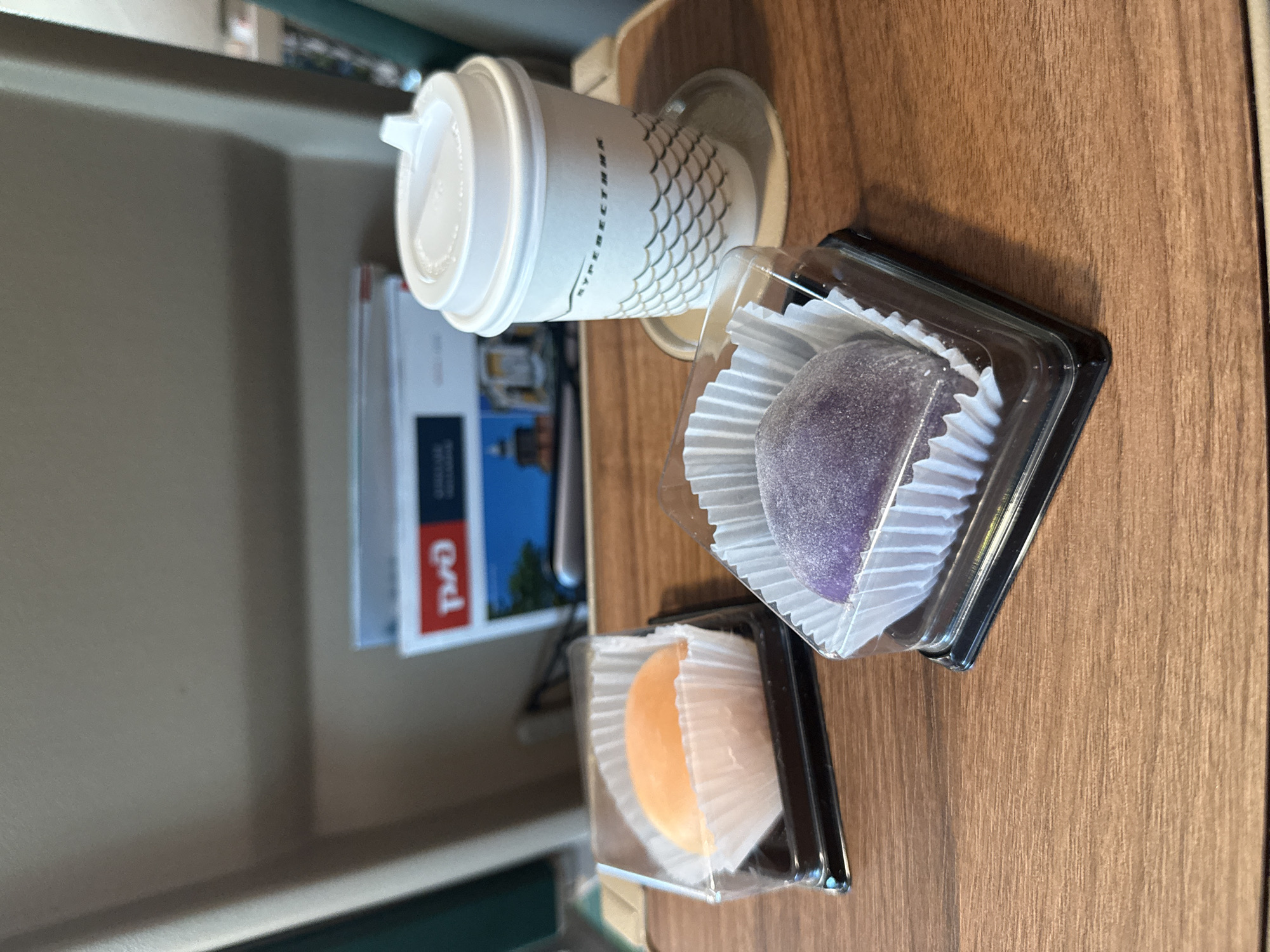 On a wooden tray, two mochi, one orange and one purple, sit in clear plastic containers. To the right, a white paper coffee cup with a lid rests on a saucer. In the background, a magazine with "РЖД" logo is visible. The scene appears to be in a train or airplane cabin given the seating and tray table. The image was taken in Nizhny Novgorod, Russia.