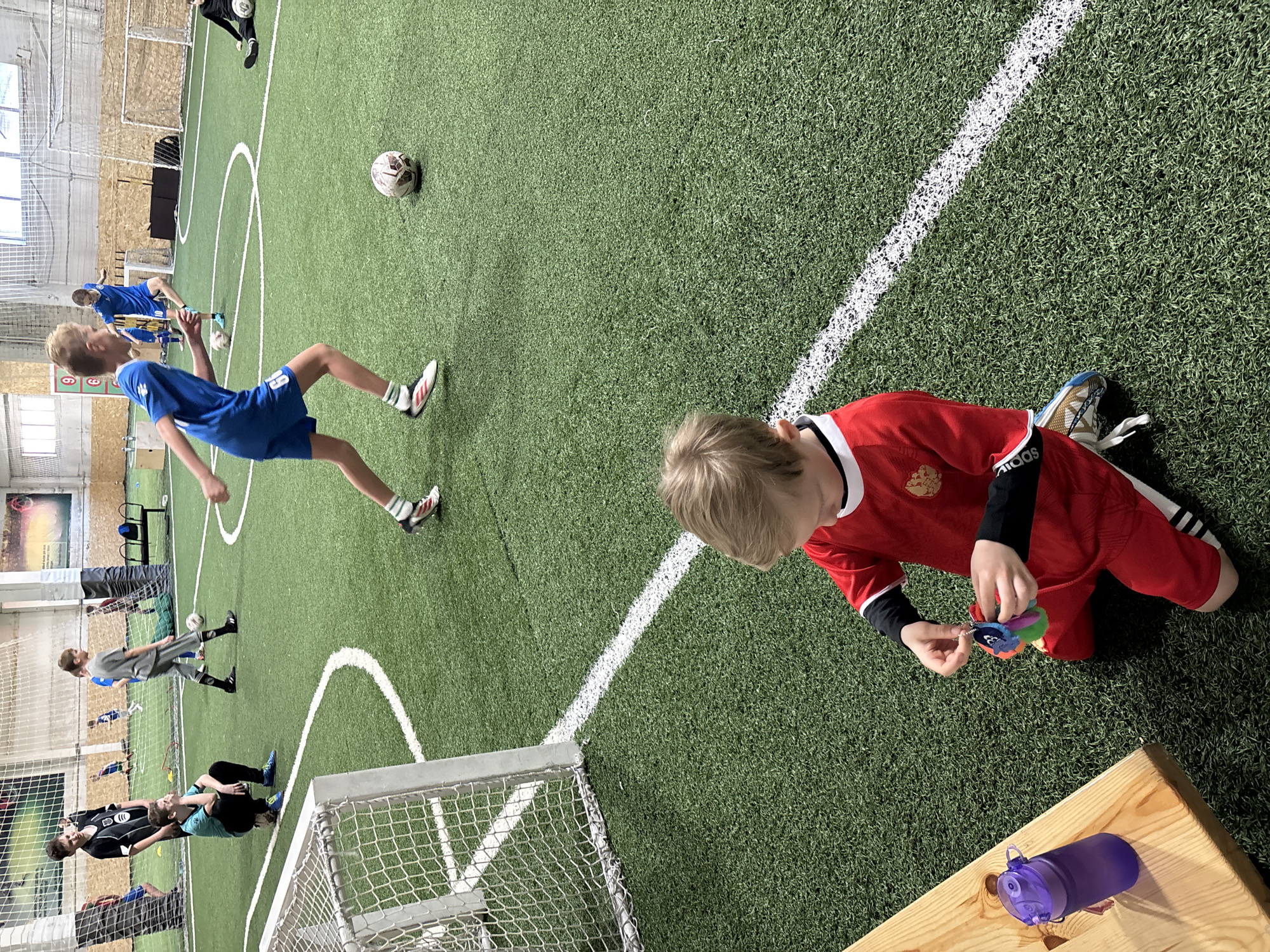 The scene is indoors, likely a sports hall or indoor soccer field in Chaikovsky, Russia. Several young people are present; some appear to be actively playing soccer, visible with balls and in athletic poses in soccer uniforms. A young person in a red soccer uniform kneels near a wooden bench, appearing to be focused on playing with an object. A purple water bottle is nearby on the bench. The playing surface is artificial turf marked with white lines indicating the soccer field boundaries. A soccer goal is in the left foreground. The building structure includes windows that provide natural light, giving evidence that the scene is indoors.