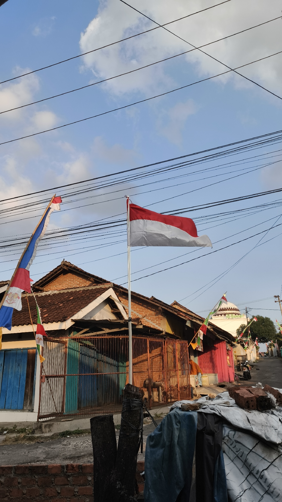 The image captures a street scene in Bandar Lampung, Indonesia, on a clear day with a blue sky and scattered clouds. Multiple power lines crisscross the upper portion of the frame. In the foreground, a rustic wooden fence and a pile of bricks are visible. Dominating the mid-ground is a building with a tiled roof, featuring red and blue doors and a rusty metal fence. Indonesian flags are prominently displayed, with a large red and white flag on a flagpole in front of the building and smaller flags adorning a tall pole to its left. In the background, a building with a domed roof, possibly a mosque, is visible, along with other structures and a street that slopes upwards. A person in an orange headscarf is standing near the pink-colored building. A motorcycle is parked on the right side of the street. The overall impression is that of a residential area decorated for a national celebration.