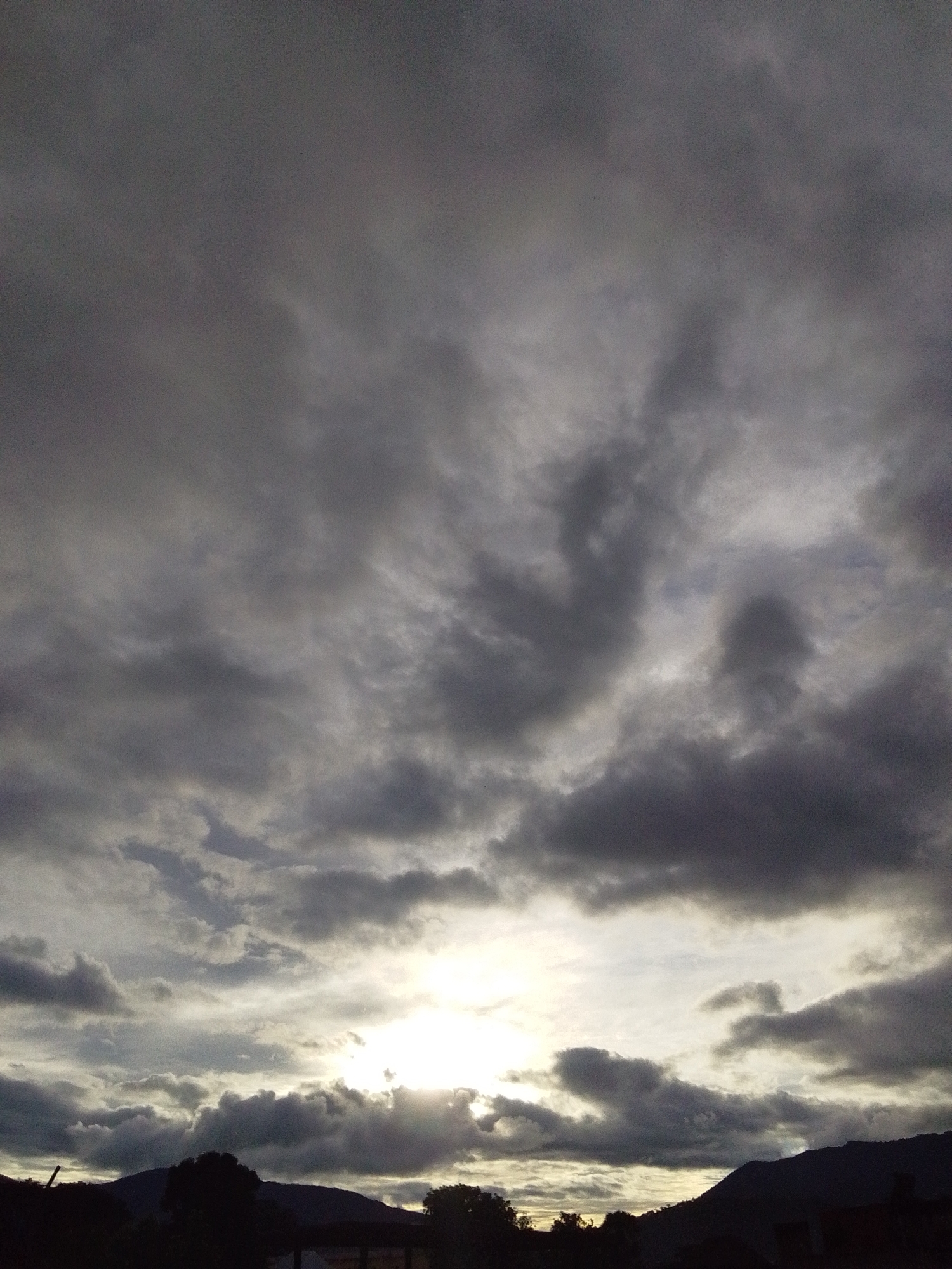 The image depicts a dramatic sky over Serti, Nigeria, with the sun partially obscured by heavy, dark clouds. The light from the sun creates a bright, diffused glow in the center of the frame, illuminating the undersides of the clouds. Silhouetted against the sky are the dark shapes of mountains and trees in the foreground, with hints of structures that may be buildings. The overall impression is one of an approaching storm or the end of a rainy day. The scene is devoid of any visible people or specific activities, focusing entirely on the natural elements and the atmospheric conditions.