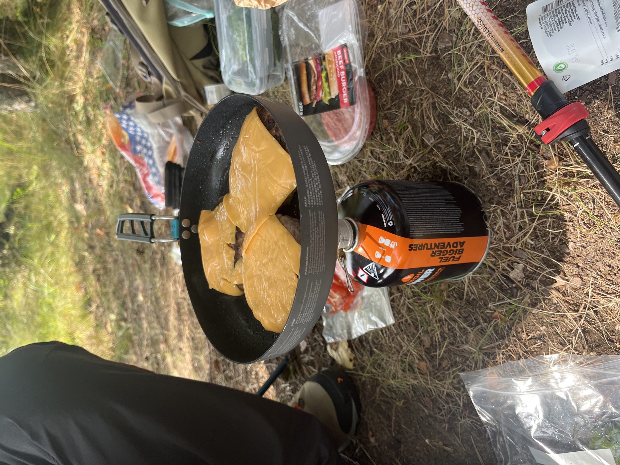 An outdoor scene depicts a cooking setup on the ground in the city of Pillapalu, Estonia. A black frying pan rests on top of an orange and black fuel canister. Inside the pan, two burger patties are being cooked, each topped with a slice of melted yellow cheese. To the right of the pan, a plastic container holds a raw burger patty and a packaged beef burger with various toppings. Further to the right, a clear plastic container with green vegetables is visible. In the background, a backpack lies on the ground, alongside a folded blanket or tarp with a red, white, and blue pattern. The ground is covered in dry grass and pine needles, suggesting a forest or wooded area. Part of a person's dark clothing and a hiking boot are visible on the left side of the image. A white plastic bag with some contents is in the lower left corner, and a section of a trekking pole with red and black accents is visible in the lower right corner.