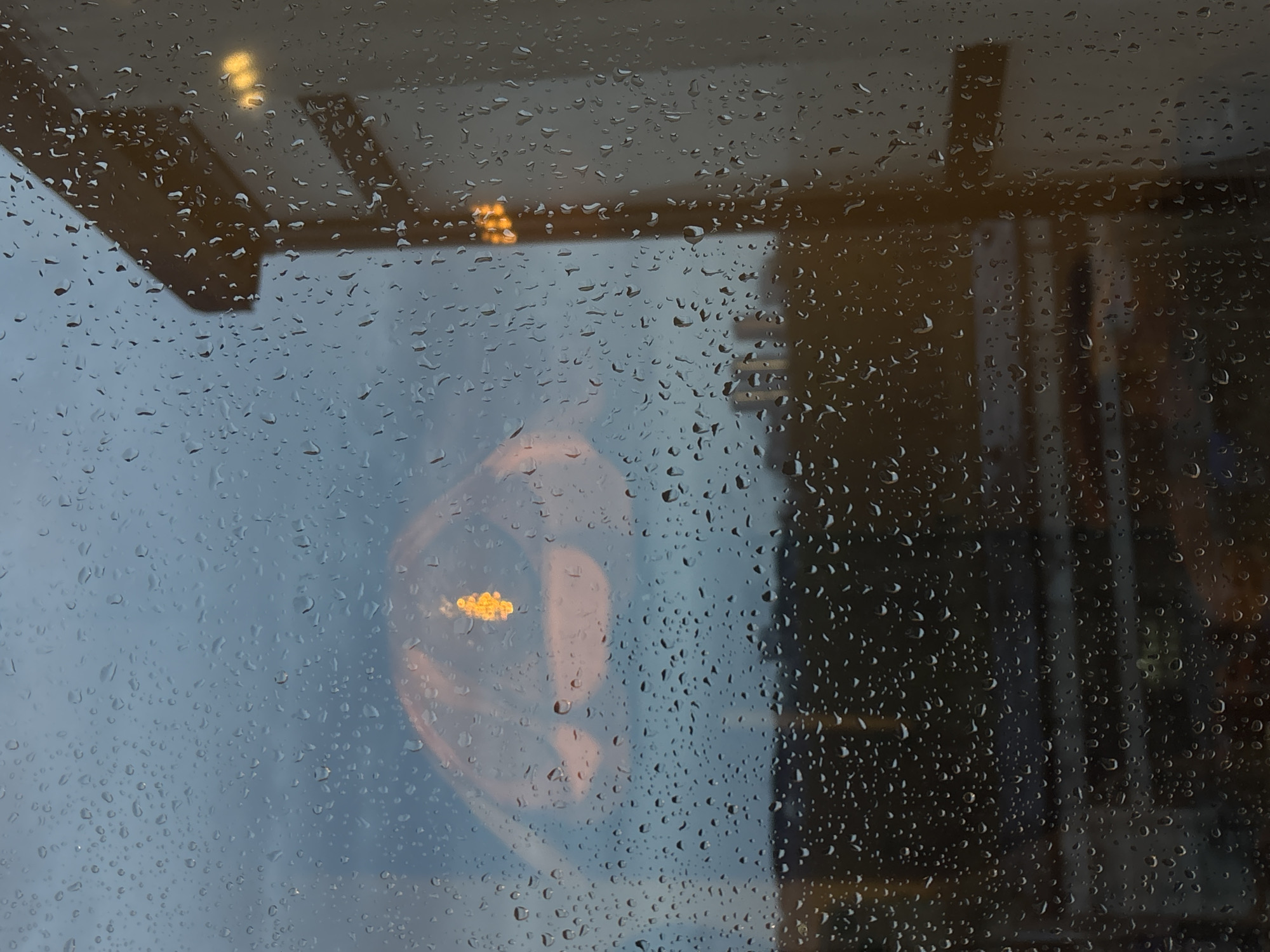 The image depicts a rain-covered window. A building interior is visible through the window, reflecting light. There is a faint image of a face superimposed over the interior scene. The location in the city of Krasnoyarsk, Russia, cannot be determined from the image.