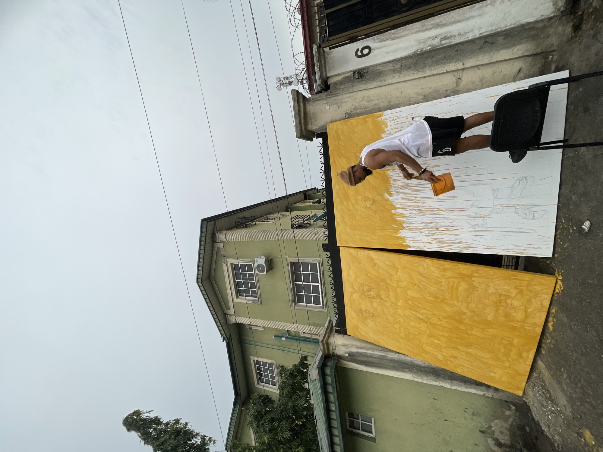 A man is painting two adjacent panels propped up on a chair in an urban setting. The panels are partially painted in a yellow color, with a sketched outline visible on the unpainted portion of the upper panel. The man wears a white tank top, dark shorts, and a light-colored head covering. The scene is located next to a building with green siding and white-framed windows. The number "6" is visible on a ledge or structure above the panels, possibly a building or gate. Barbed wire tops the gate. Power lines run overhead. The sky is overcast. This is taking place in Ado, Nigeria.