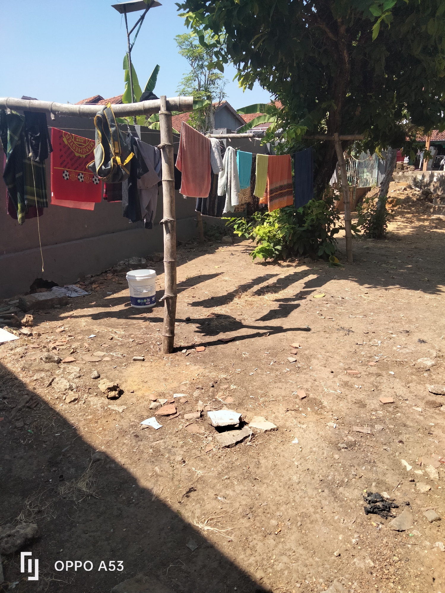 In an outdoor, sunlit area, likely a backyard in Badung, Indonesia, a clothesline made of bamboo is strung between two posts. Several articles of clothing, including shirts, towels, and what appears to be a backpack, are hanging on the line, drying in the sun. The ground is covered with dry dirt, scattered rocks, and some debris. In the background, a wall, a fence, and parts of houses are visible. A large tree with dense foliage casts shadows on the ground. A white bucket is positioned near the base of one of the clothesline posts. The sky is clear and blue.