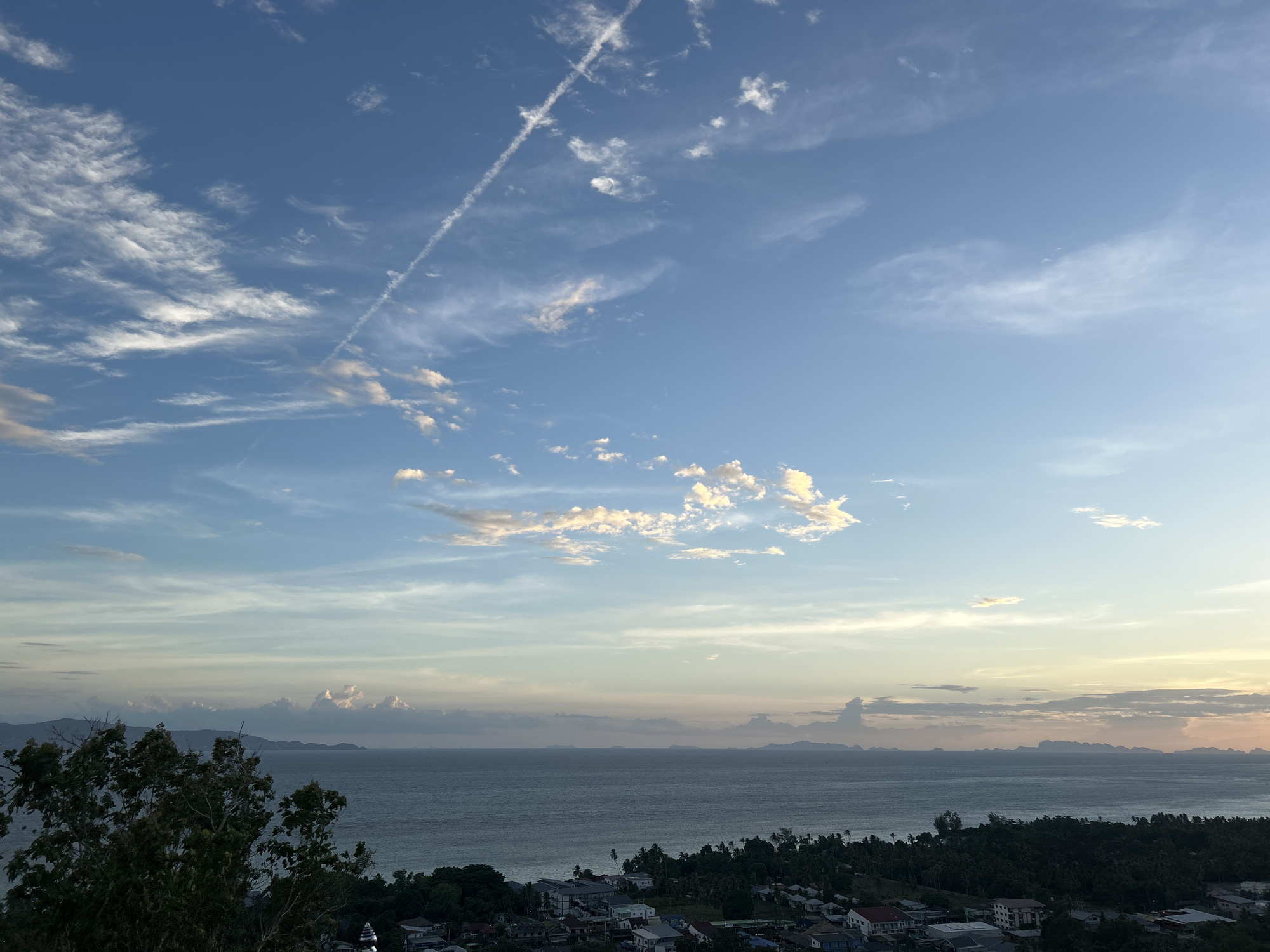The image is a scenic landscape view from a high vantage point overlooking a coastal town on the island of Ko Pha-ngan, Thailand. The sky dominates the upper portion of the frame, displaying a gradient of blue from clear overhead to softer hues near the horizon. Wispy white clouds are scattered across the sky, with a distinct contrail from an aircraft streaking diagonally from the upper left to the mid-right.
Below the sky, a wide expanse of the ocean stretches horizontally, appearing calm with gentle waves. In the distance, a series of islands or mountain formations are visible on the horizon, silhouetted against the lighter sky.
The foreground and mid-ground depict a densely populated coastal area. A dark, leafy tree occupies the far left, partially obscuring the view. Below the tree, the town is characterized by a cluster of buildings with various roof colors, including red, white, and grey. Many structures appear to be residential houses and smaller commercial buildings, interspersed with lush green vegetation and palm trees. The overall impression is of a tropical coastal settlement bathed in the soft light of either early morning or late afternoon.