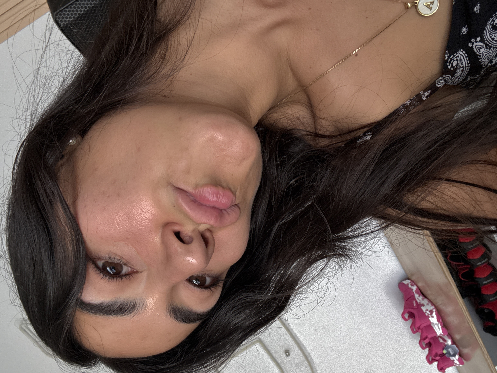 A young woman with long, dark brown hair is looking directly at the camera with slightly pursed lips. She has brown eyes and dark eyebrows. She is wearing a gold necklace with a round pendant. Her clothing is partially visible, with a black and white bandana-like pattern. The background is a plain white surface. A pink and white object is in the lower right corner of the frame. The image appears to be taken indoors with artificial lighting. There is no indication of the image being taken in Puebla, Mexico.
