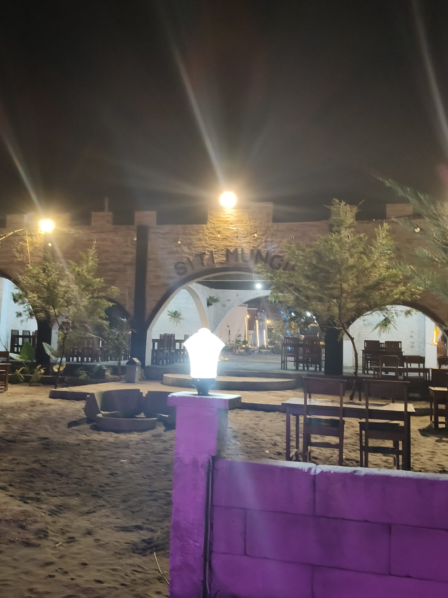 The image depicts an outdoor seating area at night, likely a restaurant or cafe, with the text "SITA MUNGIL" visible on a stone archway. The area is paved with sand and features numerous wooden tables and chairs arranged in sections. Several trees and plants are interspersed throughout the space, with some illuminated by overhead lights. A prominent modern lantern with a bright white light stands on a purple-colored stone base in the foreground. The architecture of the archway has a castle-like design with crenellations. The overall atmosphere is dimly lit, suggesting a tranquil evening setting in Desa Wonoharjo, Indonesia.