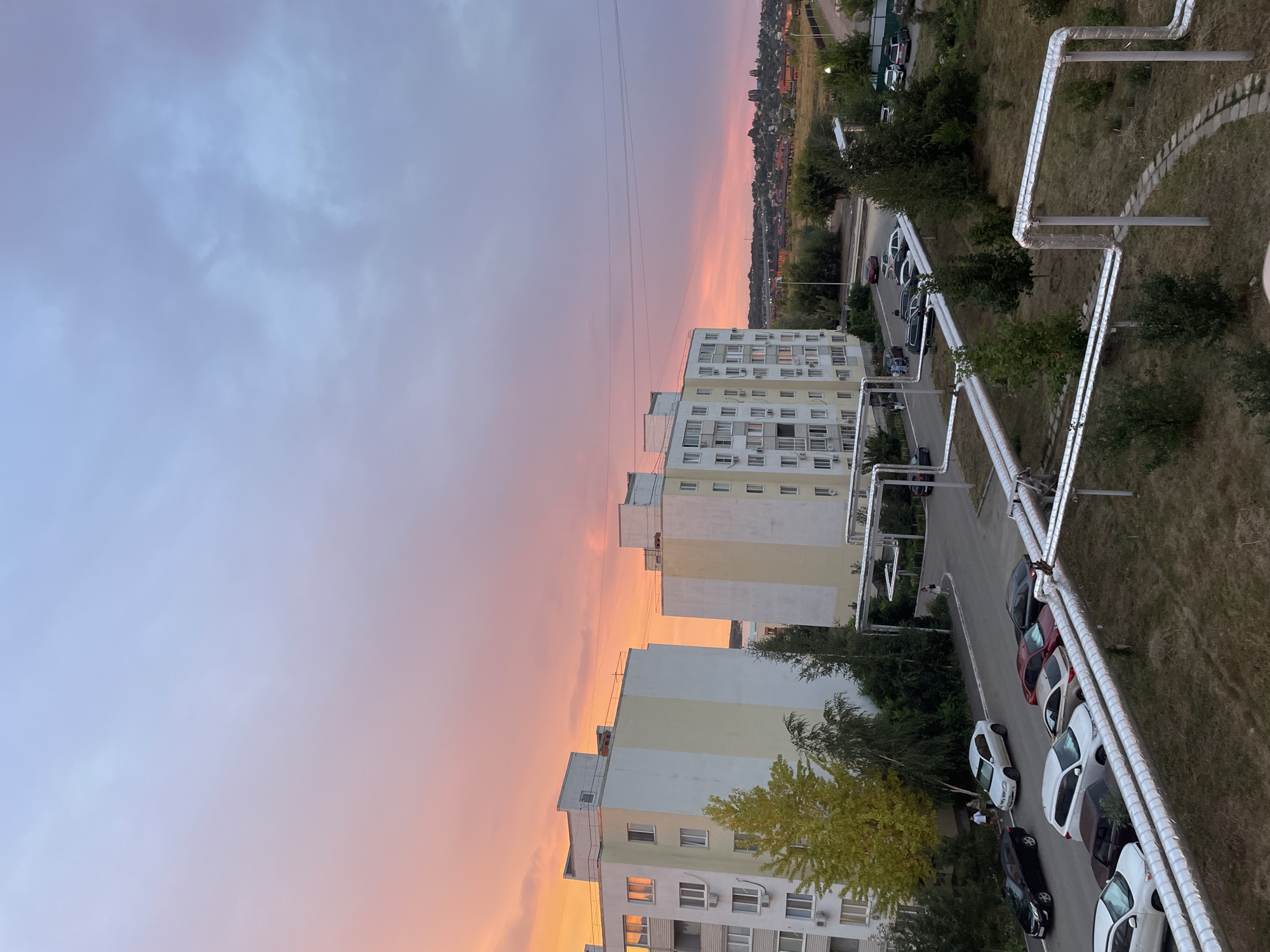 The image shows a view from an apartment building overlooking a street and neighboring buildings in Saratov, Russia. The sky is a vibrant sunset with hues of orange, pink, and blue. Several parked cars are visible on the street, as well as a few pedestrians. The foreground features large, insulated pipes running along the ground. Trees and grass are also present in the scene.