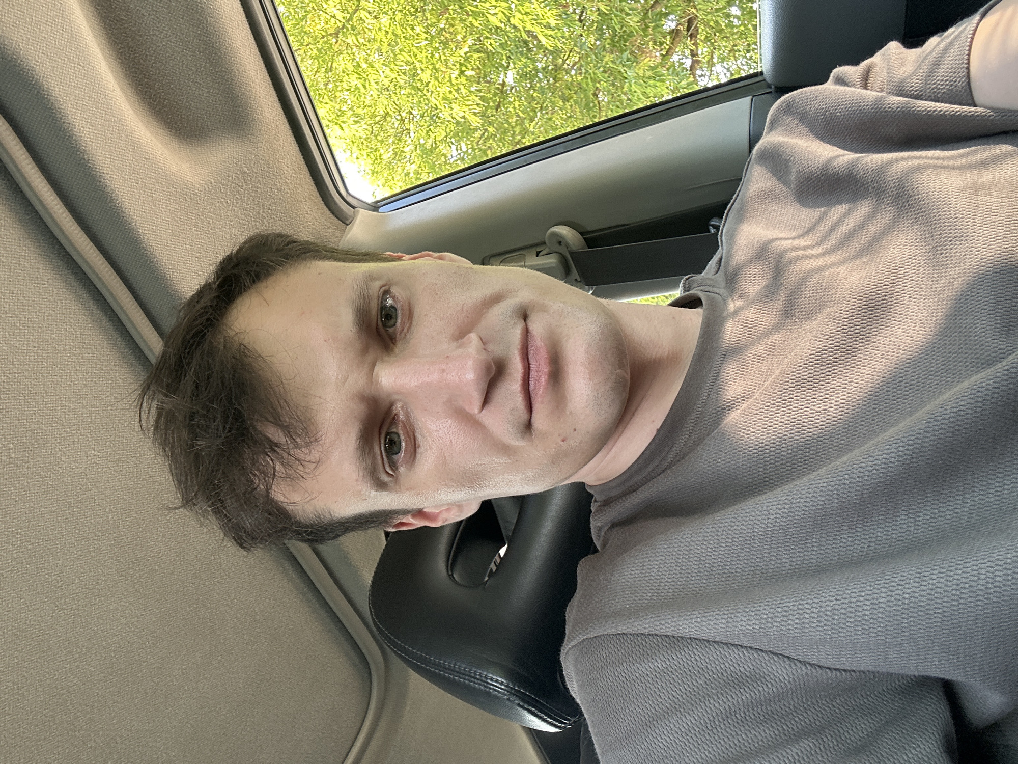 A young adult male with short, dark brown hair and fair skin is photographed from the chest up, seated inside a car. He is wearing a grey, waffle-knit crew neck shirt. The car's interior, including the headrest and the car's roof lining, is visible. Sunlight filters through the trees outside the passenger window, casting dappled shadows on the man and the car's interior. The background shows lush green foliage, suggesting a natural, outdoor setting. The image was taken at an unknown location within the city of Makiv, Ukraine.