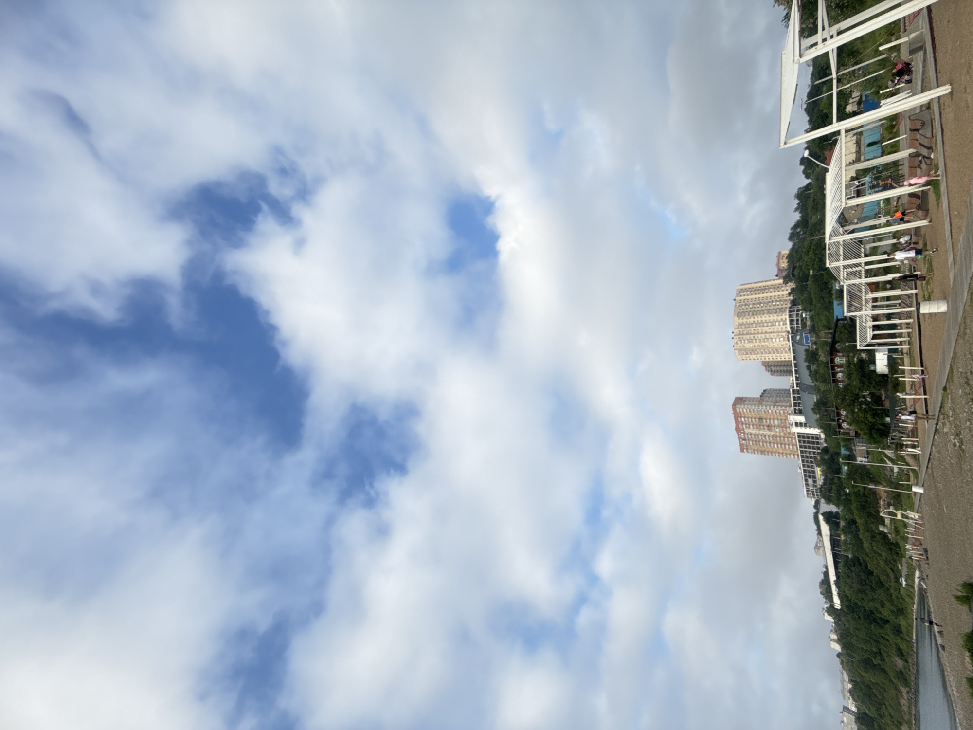 The image captures a coastal scene in Vladivostok, Russia, likely from a beach or promenade area. In the foreground, a gravelly beach slopes down to the water. Scattered across the beach are various outdoor amenities, including a white, open-air structure with multiple sections, possibly a rest area or playground, with some individuals visible near it. Further back, a green, tree-covered slope rises, culminating in a cluster of multi-story buildings on the hilltop, characteristic of urban development on uneven terrain. The sky above is a mix of blue sky and scattered white clouds.