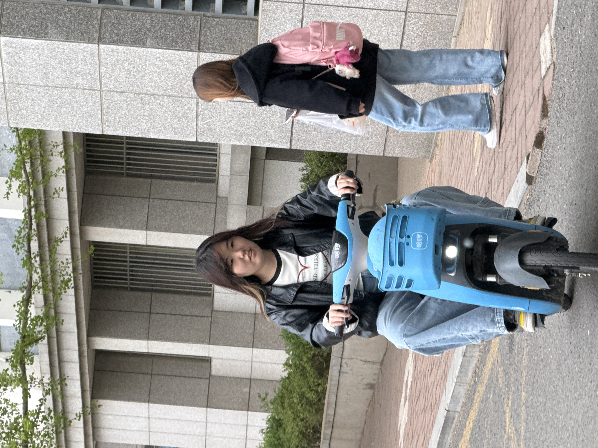 The image shows two young women in an urban setting, likely in Zhongtun, China, during the daytime under bright, possibly overcast, conditions.
In the foreground, a woman with long, dark hair is seated on a bright blue shared bicycle or scooter, gripping the handlebars. She wears a black leather-like jacket over a white t-shirt with black trim and partially visible text, "ND THES" and "R LEARI". She is also wearing blue denim jeans and light-colored sneakers, and looks towards the camera with a neutral expression. The blue vehicle has a basket-like front section with a visible headlight and Chinese characters, likely "哈啰" (HelloBike), a popular shared bicycle service.
Behind her, slightly further back on a paved sidewalk, another woman is walking away from the camera. She has long brown hair and is dressed in a black hooded jacket, light blue denim jeans, and white sneakers. She carries a pink backpack adorned with a white plush keychain and a pink one. A white plastic bag is also visible hanging from her left hand.
The background features a modern building with a facade composed of grey and white rectangular tiles. To the left, some green foliage from bushes and a small tree adds a touch of nature. The ground consists of concrete paving stones on the sidewalk and asphalt road adjacent to it, with some faint yellow road markings.