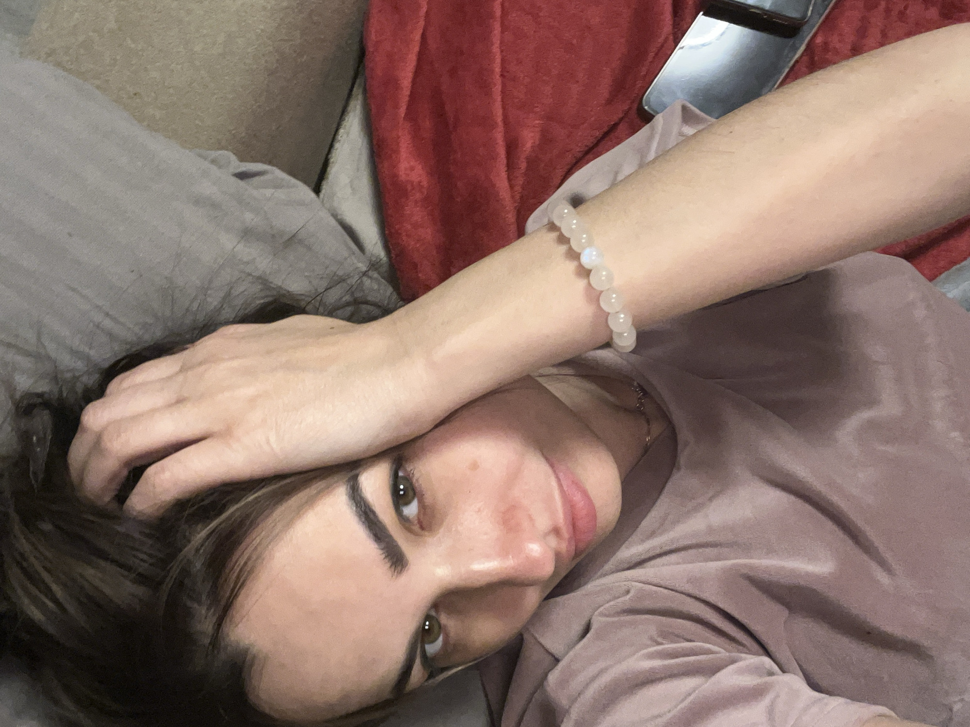 A woman is lying on a gray pillow next to a red blanket. She is wearing a pink shirt and a bracelet made of white beads. Her right hand is above her head, resting on her hair. A cell phone is visible next to her arm on the right. The location and city are not discernable from the image.