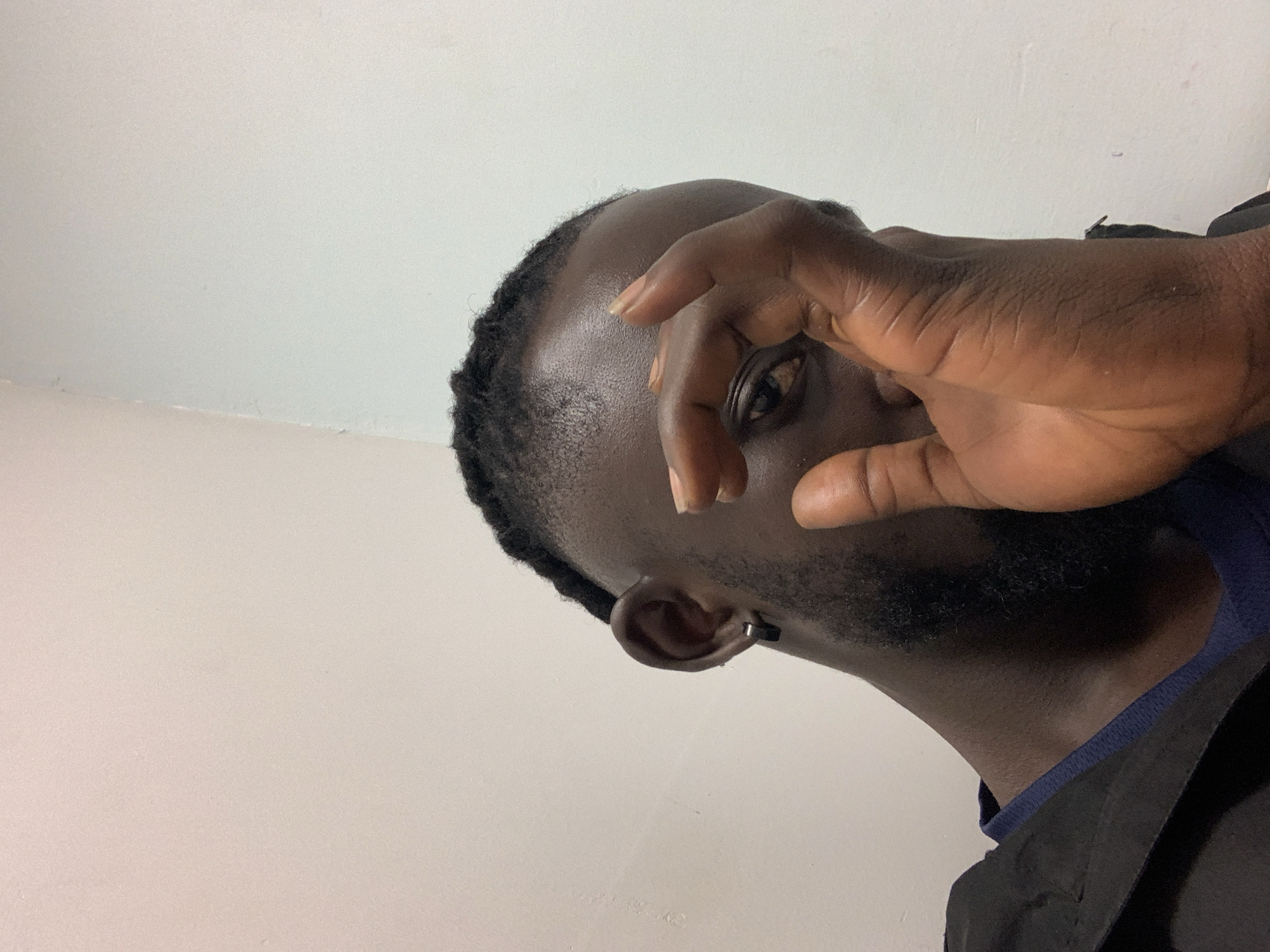 A close-up, low-angle, upward-tilted shot features a dark-skinned man with a beard, partially obscuring his face with his hand in a circular gesture, leaving one eye visible. He has an earring in his left ear. The background is a plain white or light-colored wall. There is no evidence to determine if this image was taken in Lekki, Nigeria, or anywhere else.