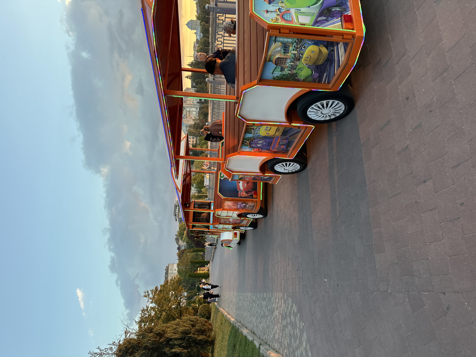 In Ternopil, Ukraine, a decorative tourist train with cartoon characters on its side is parked along a paved walkway. The train's carriages are adorned with colorful depictions of characters from the animated series "Tayo the Little Bus" and other animated characters. The walkway is bordered by a grassy area with trees on the left and a body of water with a railing on the right. In the background, buildings and a domed structure are visible under a partly cloudy sky during what appears to be sunset or early evening. Several people are walking on the walkway and seated on the train. The scene is tranquil and suggests a public recreational area.