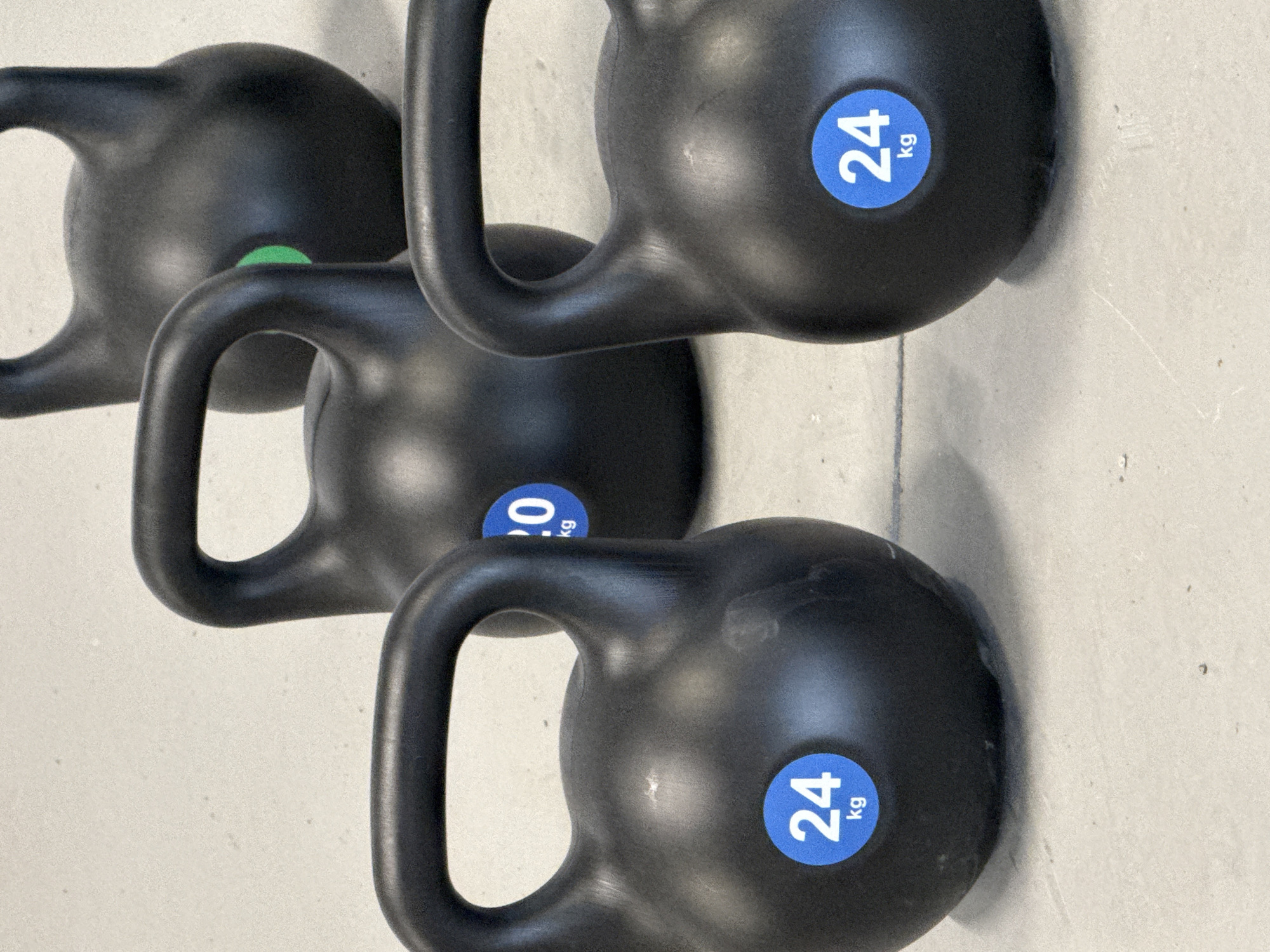 The image shows a close-up, angled view of several black kettlebells. The kettlebells appear to be resting on a light-colored surface. One kettlebell has a green accent while the others have blue accents, with "24 kg" printed on them. There is nothing to indicate that this image was taken in Sin'kovo, Russia.