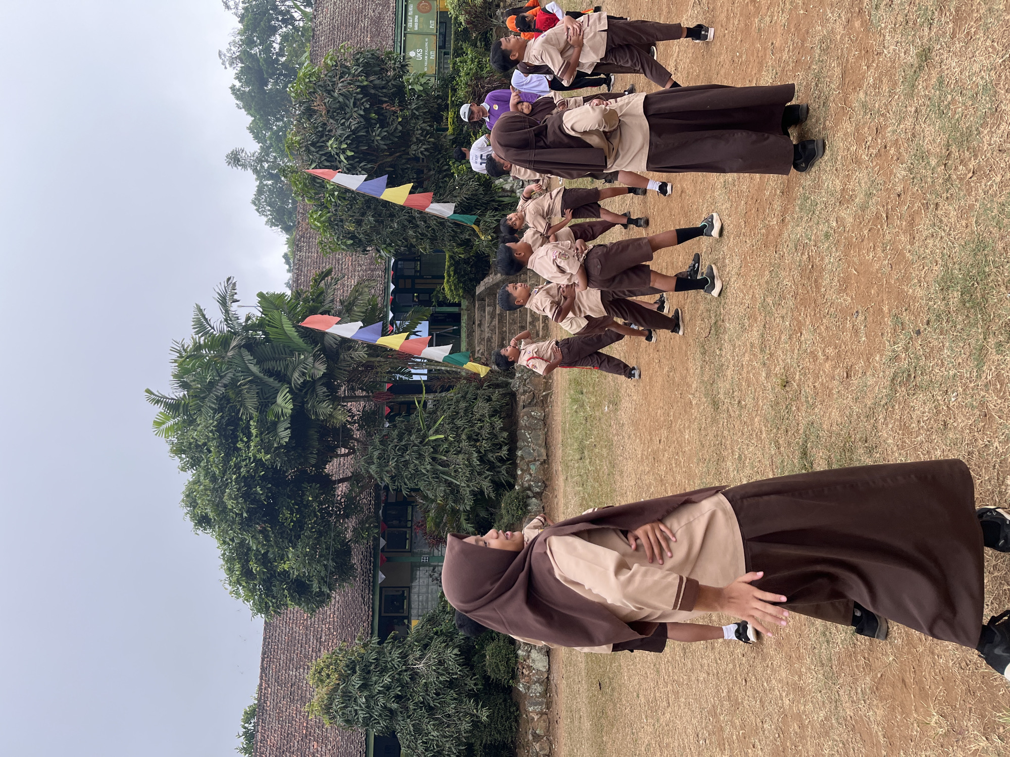 The image depicts an outdoor scene in front of a school building in Gembong, Indonesia. A group of uniformed schoolchildren, wearing brown and tan uniforms, are engaged in what appears to be a physical activity or dance routine on a dirt field. Some of the children are wearing headscarves. In the background, a two-story school building with red tiled roofs is visible, adorned with colorful flags. Several large trees and lush greenery surround the building. A few adults are also present, one of whom appears to be supervising the children. The sky is overcast, suggesting a cloudy day. The overall scene conveys a sense of communal activity and education.