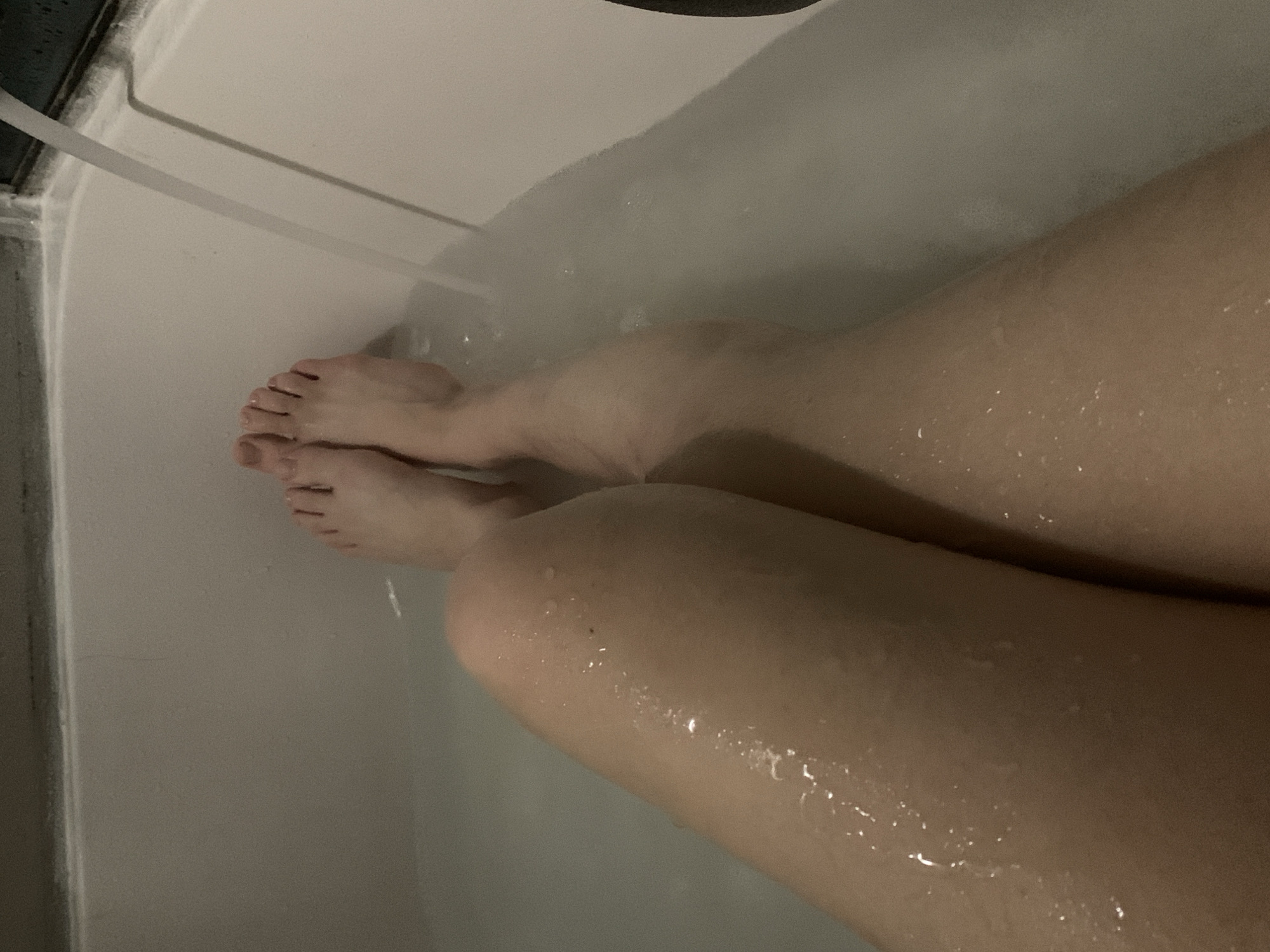 A person is sitting in a bathtub. The person's legs are visible from the knees down, and their feet are near the edge of the tub. Water is running into the tub. The location is not discernible from the image.