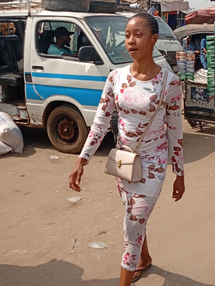 A young woman in a floral dress and beige purse strides down a street. A van with a driver inside is parked nearby, with bags piled on its roof. A loaded cart and another vehicle are visible in the background. The street is littered with trash. The woman is the central focus, suggesting a candid shot in a busy, perhaps outdoor marketplace.