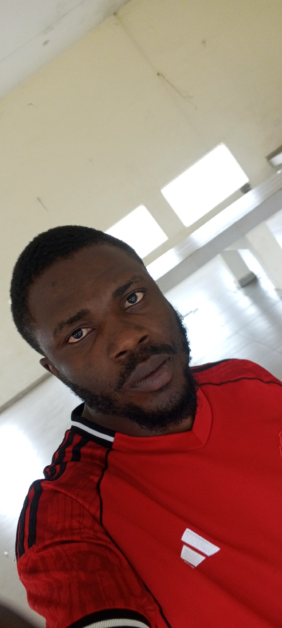 The image is a selfie of a Black male in his late 20s or early 30s, looking directly at the camera. He has dark skin, a close-cropped haircut, and a short beard. He is wearing a red t-shirt with black stripes on the sleeves and a white logo on the chest. The background appears to be an indoor space with a ceiling with recessed lighting. The lighting suggests it might be a public building such as a school or office, and the lighting fixtures are indicative of fluorescent or LED panel lights. The floor appears to be tiled or polished concrete. The image was taken in Jos, Nigeria.