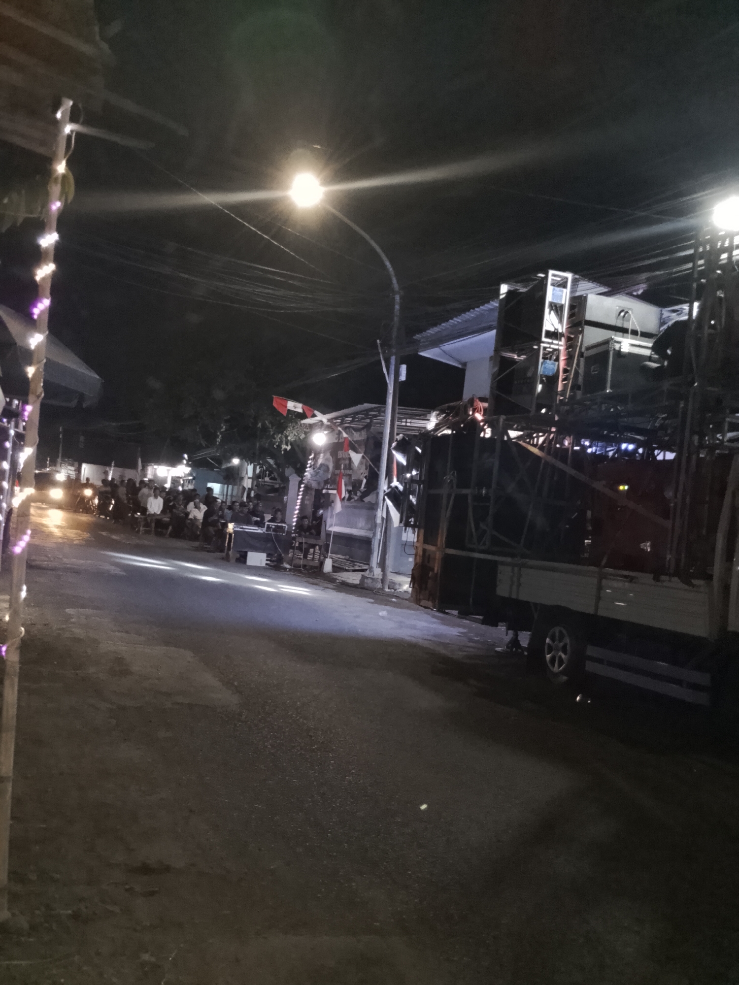This is a nighttime photograph taken on a street in Pasuruan, Indonesia. The scene depicts an outdoor gathering, possibly a local event or performance, given the presence of a large sound system mounted on a truck. The truck is heavily laden with speakers and stage lighting equipment, suggesting it is set up for a performance.
In the foreground, the asphalt road is visible, with some reflections from streetlights and potentially other light sources. To the left, there is a narrow strip of sidewalk or adjacent area adorned with string lights, some of which appear to be purple and white. Further down the street on the left, a group of people are seated, and there are vehicles, including motorcycles with their headlights on, indicating movement or waiting.
On the right side of the street, the large sound system on the truck dominates the view. The truck appears to be parked alongside a building or stall with a corrugated metal roof. Behind the truck, more structures and trees are visible, partially obscured by the darkness and the equipment.
A street lamp is prominent in the midground, casting a bright light and creating lens flare. Several individuals are visible in the midground, some standing and some seated, gathered around a setup that might be a DJ booth or a control station for the sound system. Flags, possibly national or event-related, are also present, attached to poles near the structures on the right. The overall atmosphere is that of an illuminated, active street scene at night, with a focus on a sound system and an assembled audience.