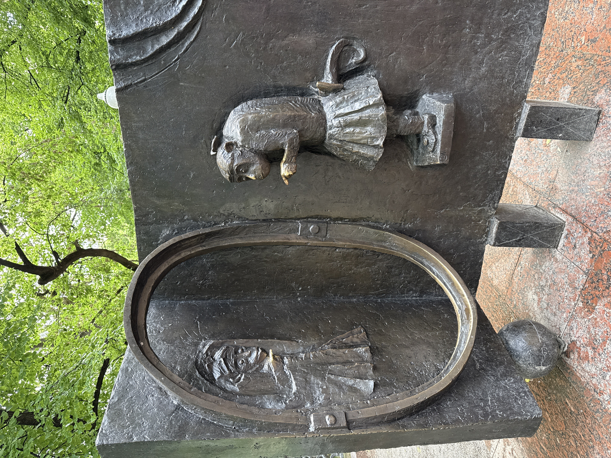 This image depicts a bronze bas-relief sculpture affixed to a dark stone monument. The sculpture shows two scenes: at the top, a figure in a dress or tutu, appearing to bow or bend forward. Below it is an oval frame enclosing the relief of a person lying down, possibly deceased. The monument stands outdoors, with foliage visible in the background and what appears to be granite siding. Based on the imagery, specifically the representations of figures, this may be a monument related to the arts. The description is in Moscow as it is the only city in the world with that name, and where it is likely this sculpture is.