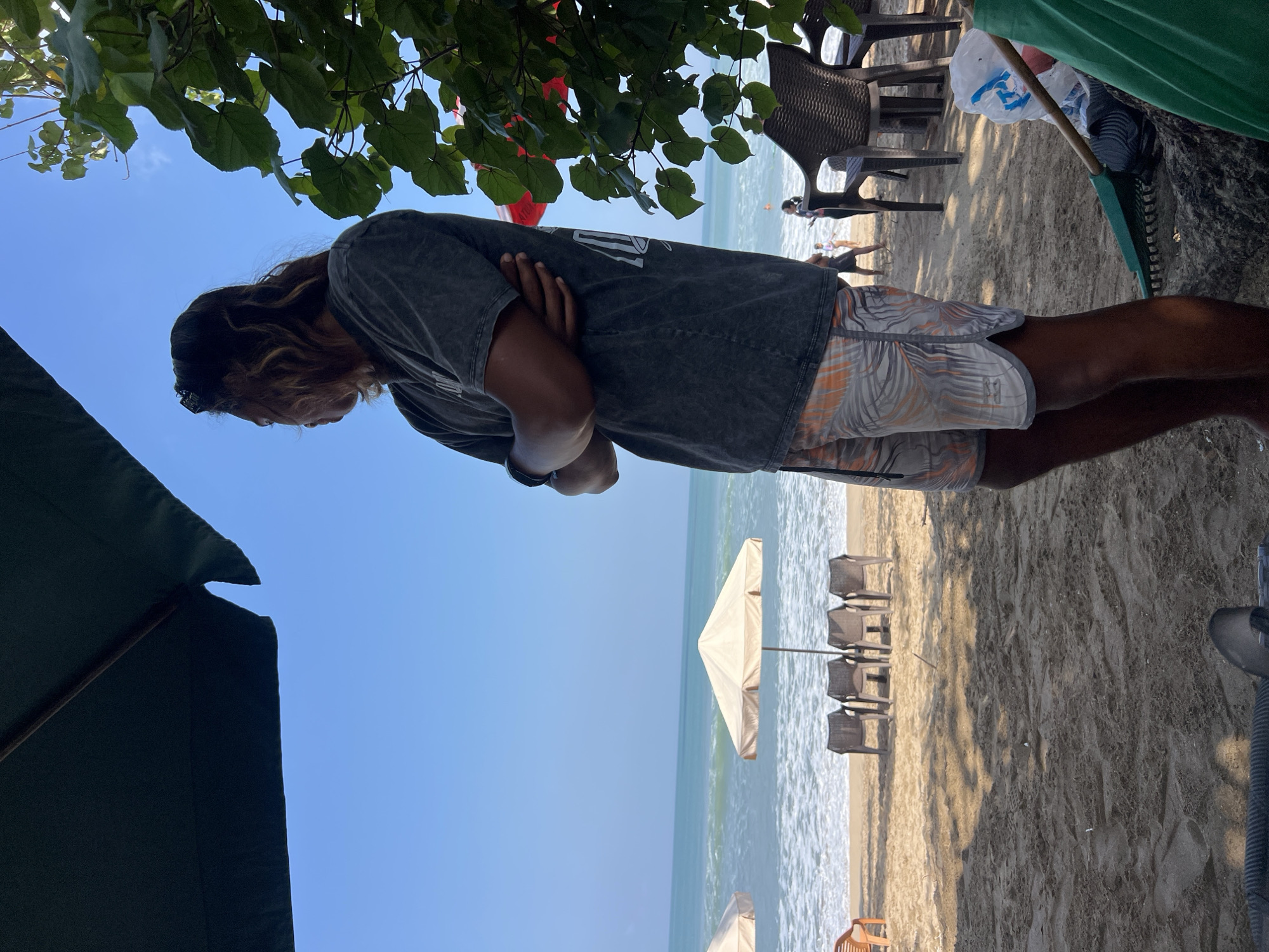 An individual stands on a sandy beach in Kuta, Indonesia, with their arms crossed, facing away from the camera and looking towards the ocean. The person is wearing a dark grey t-shirt and patterned shorts. In the background, there are beach umbrellas, chairs, and people walking on the sand. The ocean is visible with waves breaking on the shore, and the sky is clear and blue. Lush green foliage is present to the right of the frame, and a dark umbrella is positioned in the upper left corner, casting a shadow.