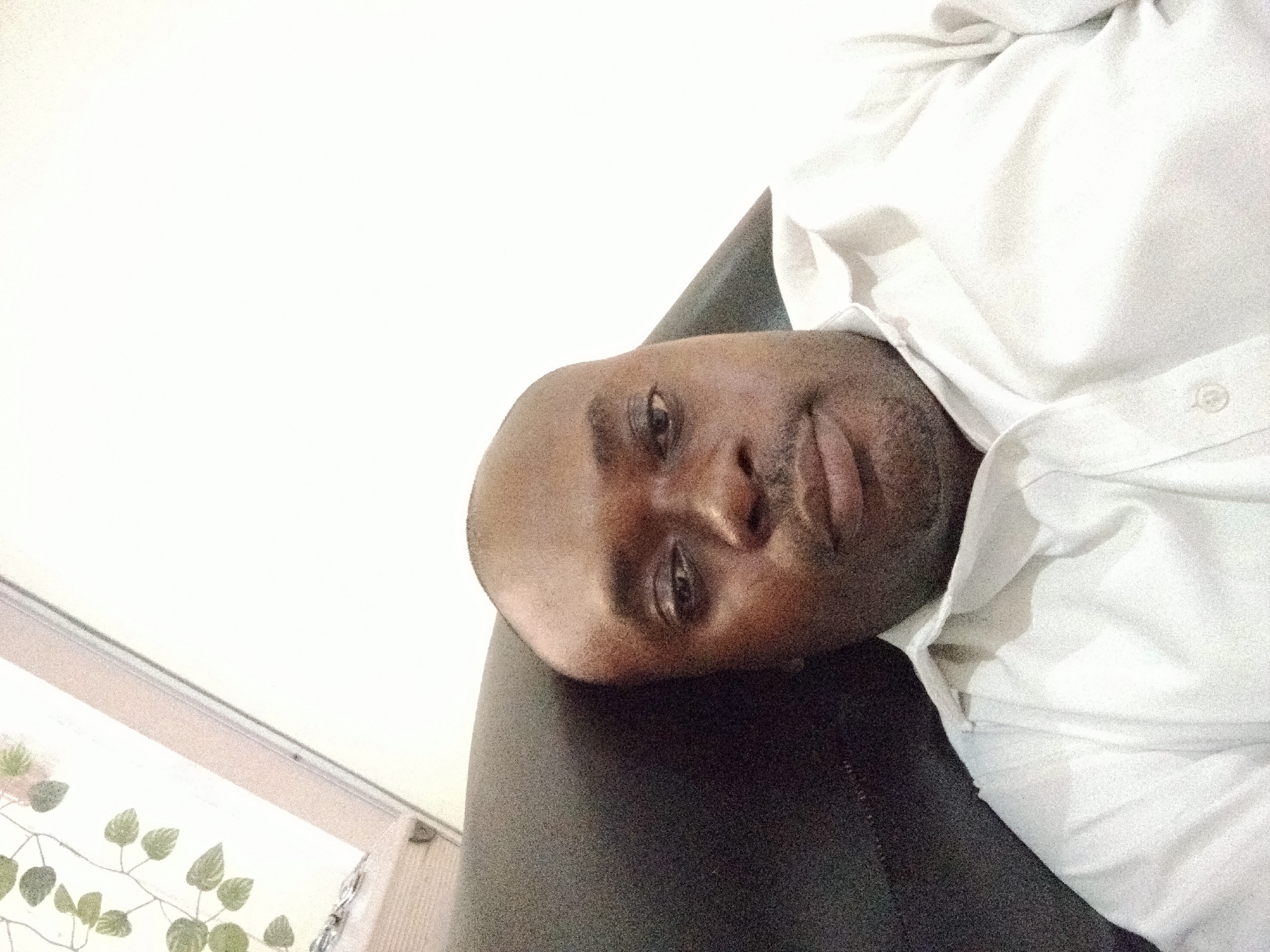 The image shows a man of dark complexion lying down, with his head resting on a dark, upholstered surface, likely a couch or bed. He is wearing a white, long-sleeved collared shirt that appears slightly rumpled. His head is tilted to his right, and he looks directly at the viewer with a relaxed, neutral expression. He has a shaved head and some facial stubble or a short beard.
The setting is indoors, characterized by bright, even lighting. A significant portion of the background is a plain, bright white wall or ceiling. In the lower left corner, a window or framed opening is partially visible, featuring a decorative element resembling a vine with green leaves, adding a touch of natural or artificial greenery to the interior space. The absence of harsh shadows suggests a uniformly lit environment, possibly during the daytime.
There is no visible text in the image. The overall scene conveys a sense of quiet and repose within an indoor setting in Gwarinpa, Nigeria.