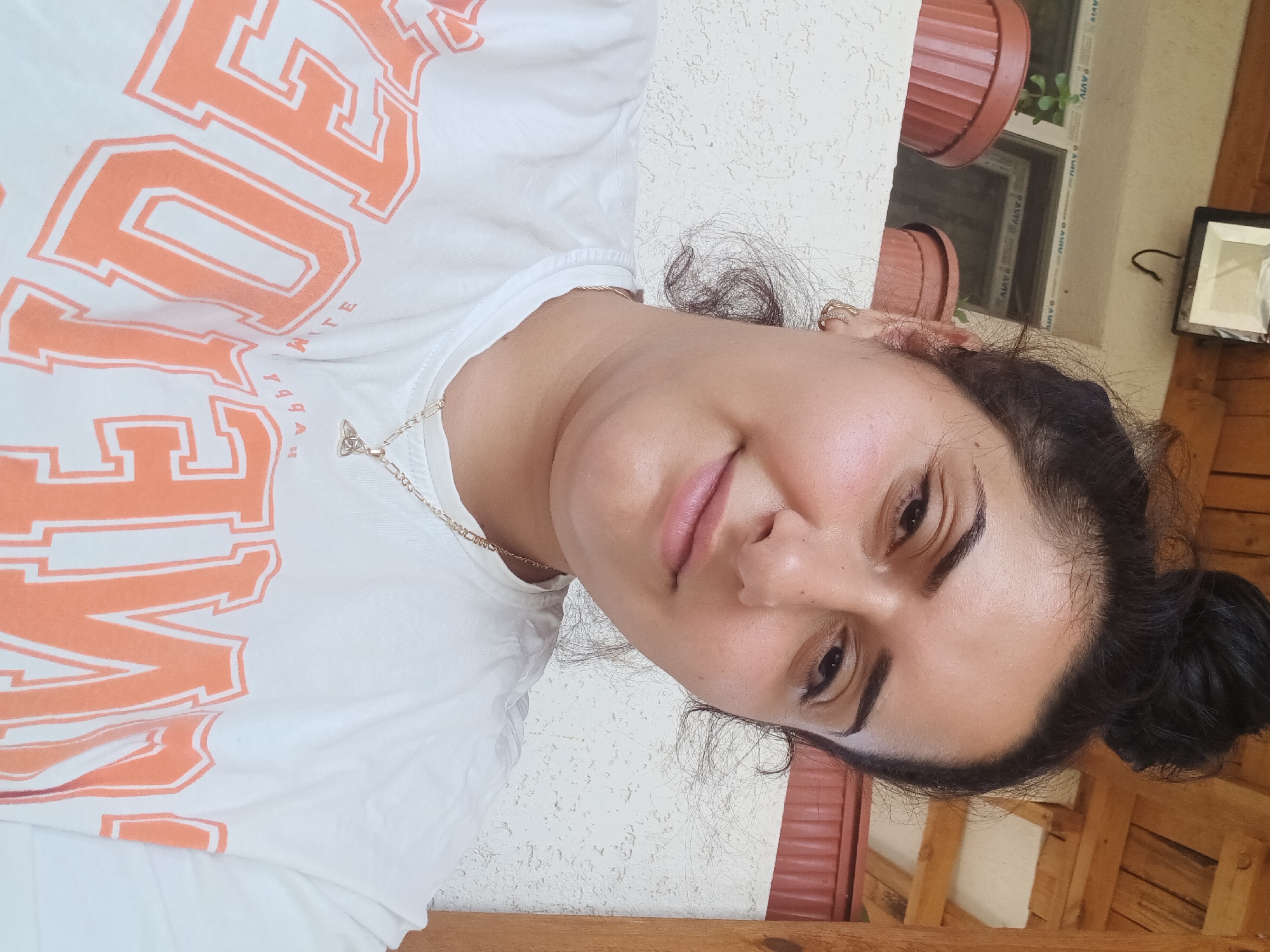 In Zimnitsa, Bulgaria, a woman with dark, curly hair styled in a bun poses for a selfie on a covered porch. She wears a white t-shirt with large orange lettering that reads "HAPPY." A delicate gold chain with a triquetra pendant rests on her neck. Behind her, a textured white wall is visible, with wooden beams and a corrugated metal roof overhead. Two brown potted plants sit on a ledge to her left, and a black outdoor light fixture is mounted on the wall above them.