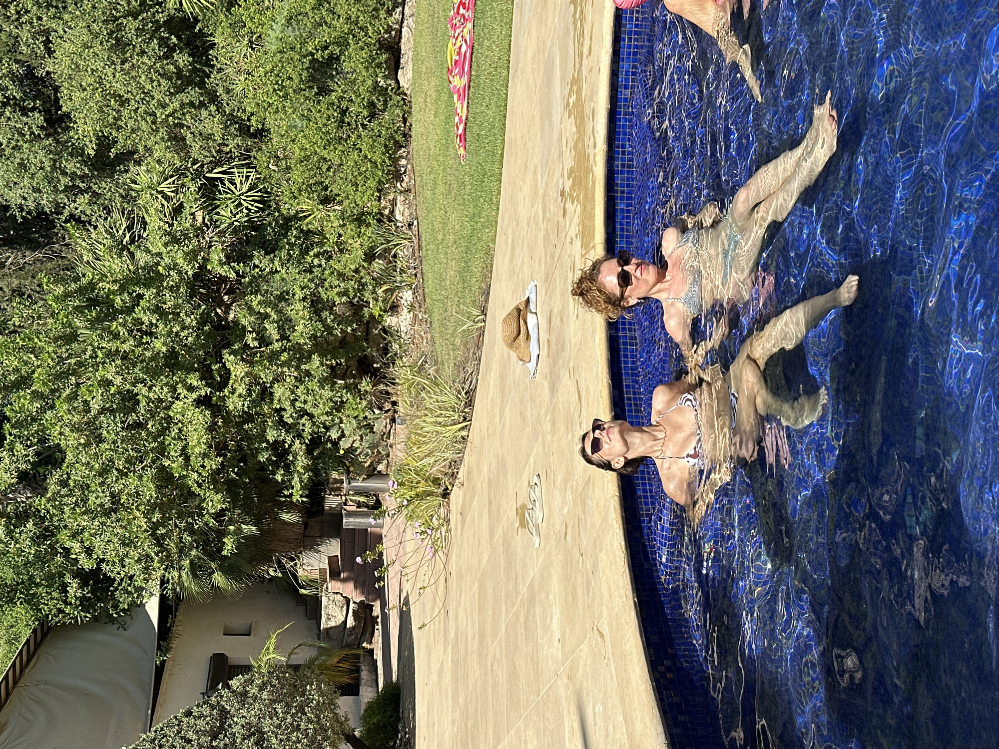 The image depicts an outdoor swimming pool scene in Savyon, Israel. In the foreground, two women are relaxing in the blue, tiled pool. The woman on the left, wearing sunglasses and a bikini with a patterned top and bottoms, is tilting her head back and closing her eyes. The woman on the right, also wearing sunglasses and a bikini, is looking to her right. Their legs are visible submerged in the water. In the background, a stone patio surrounds the pool, and beyond that, lush green vegetation and trees create a natural backdrop. A building with a light-colored awning is visible in the upper left, partially obscured by foliage. On the patio, a straw hat rests on a white towel, and a pair of flip-flops are placed nearby. A colorful beach towel lies on the grass in the far background. The overall impression is one of leisure and relaxation in a sunny, Mediterranean climate.