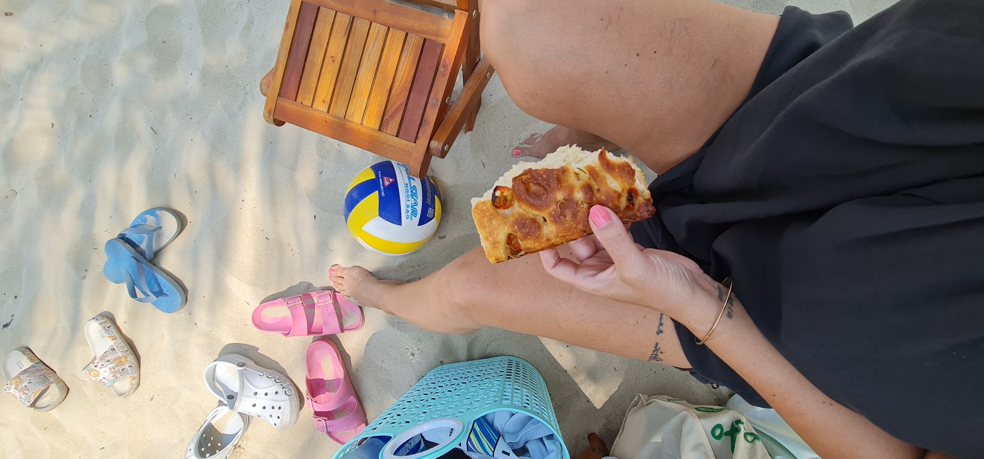The image presents a bright, top-down view of a sunny beach scene in Đà Nẵng, Vietnam. A person is seated on the light-colored sand, with their legs and one arm visible. They are wearing black clothing, likely shorts or a skirt, and have pink painted toenails and fingernails. The person is holding and eating a slice of focaccia-like bread, topped with what appear to be small tomatoes or peppers and onions.
Various items are scattered on the sand around the individual. A blue, yellow, and white volleyball, bearing the text "SWEEVE", "NGOI SAO", and "ALL STAR", rests near a small, wooden foldable beach chair. Several pairs of footwear are visible, including light blue flip-flops, pink slides, white Crocs, and patterned slides. A light blue plastic woven basket, containing some folded items, is placed next to a partially visible light green cloth or reusable bag, which has "ACTION" and possibly "OFO" printed on it. The person wears a gold-colored bangle on their wrist, and a small, indistinct tattoo is visible on their forearm. The scene conveys a relaxed beach outing under bright, clear daylight.