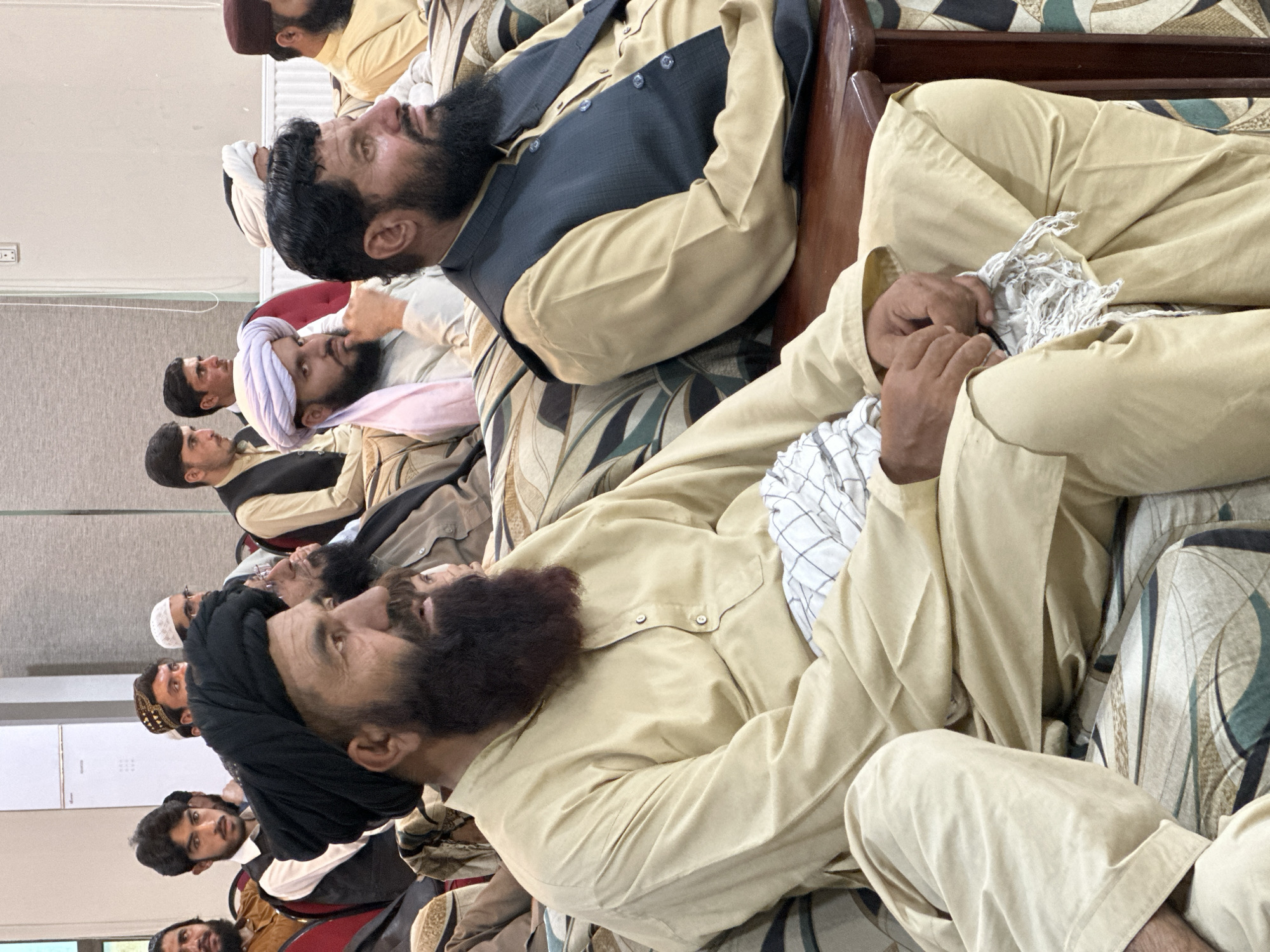 In a location identified as Quetta City Tehsil, Pakistan, a group of men, primarily clad in traditional attire consisting of long, loose-fitting beige shirts and pants, are seated close together in rows. Several men also wear head coverings, including turbans of varying colors and designs. A few men are wearing vests over their shirts. Most of the men have beards. Some men are holding and manipulating white cloths or scarves. The setting appears to be indoors, on what looks like cushioned benches.
