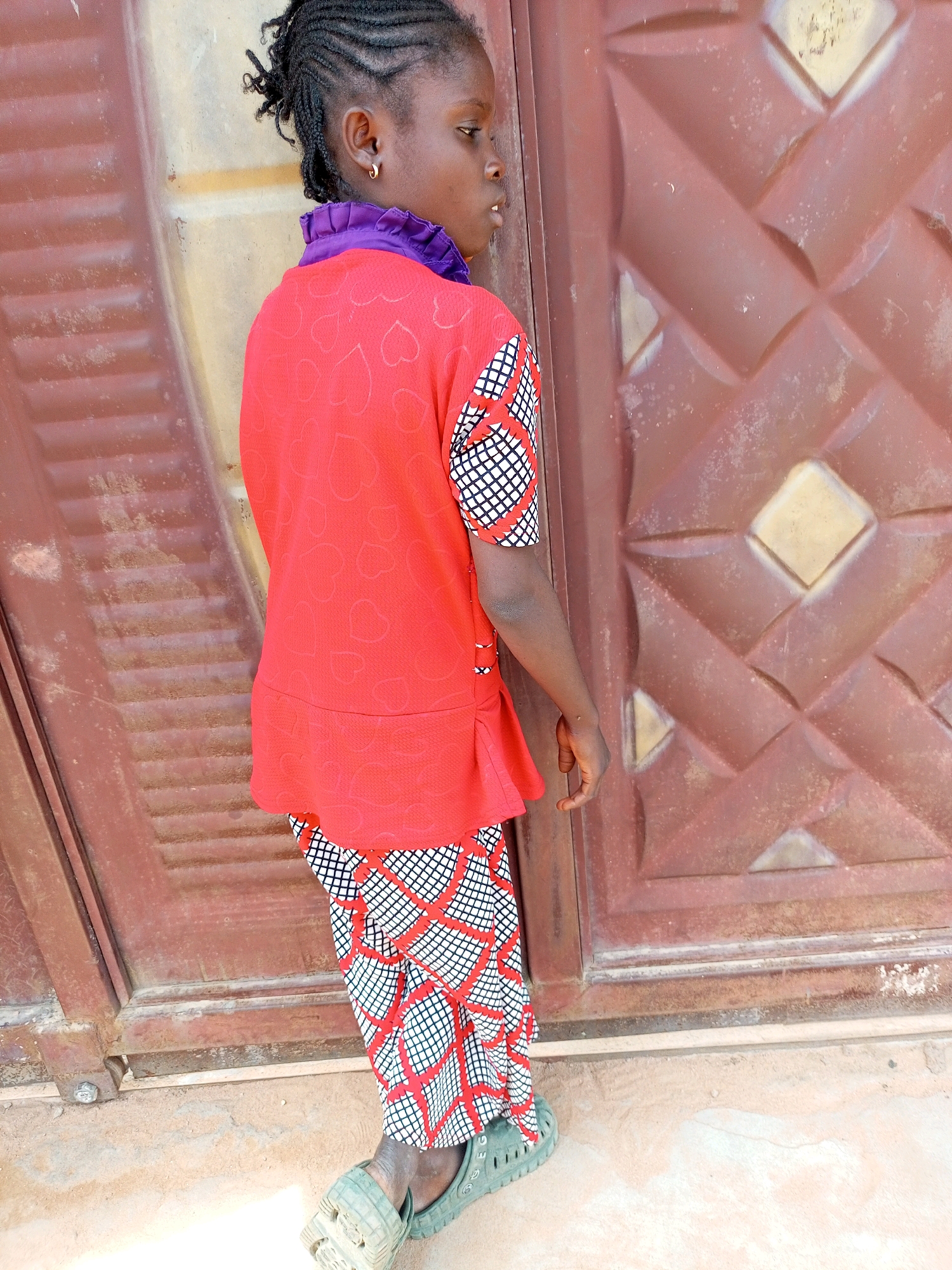 A young girl with braided hair, wearing a red and white patterned outfit with a purple ruffled collar, stands in front of a textured brown door. The girl is looking to the right, with a slight tilt of her head. She is wearing green slip-on shoes. The door is large and appears to be made of metal, with a repeating diamond pattern. The ground beneath her feet is a dusty, sandy surface. The background suggests an outdoor setting, possibly a courtyard or alleyway in the city of Tempure, Nigeria. The lighting indicates it is daytime, with a slight shadow cast on the ground. The overall scene is a candid portrait of the girl in her environment.