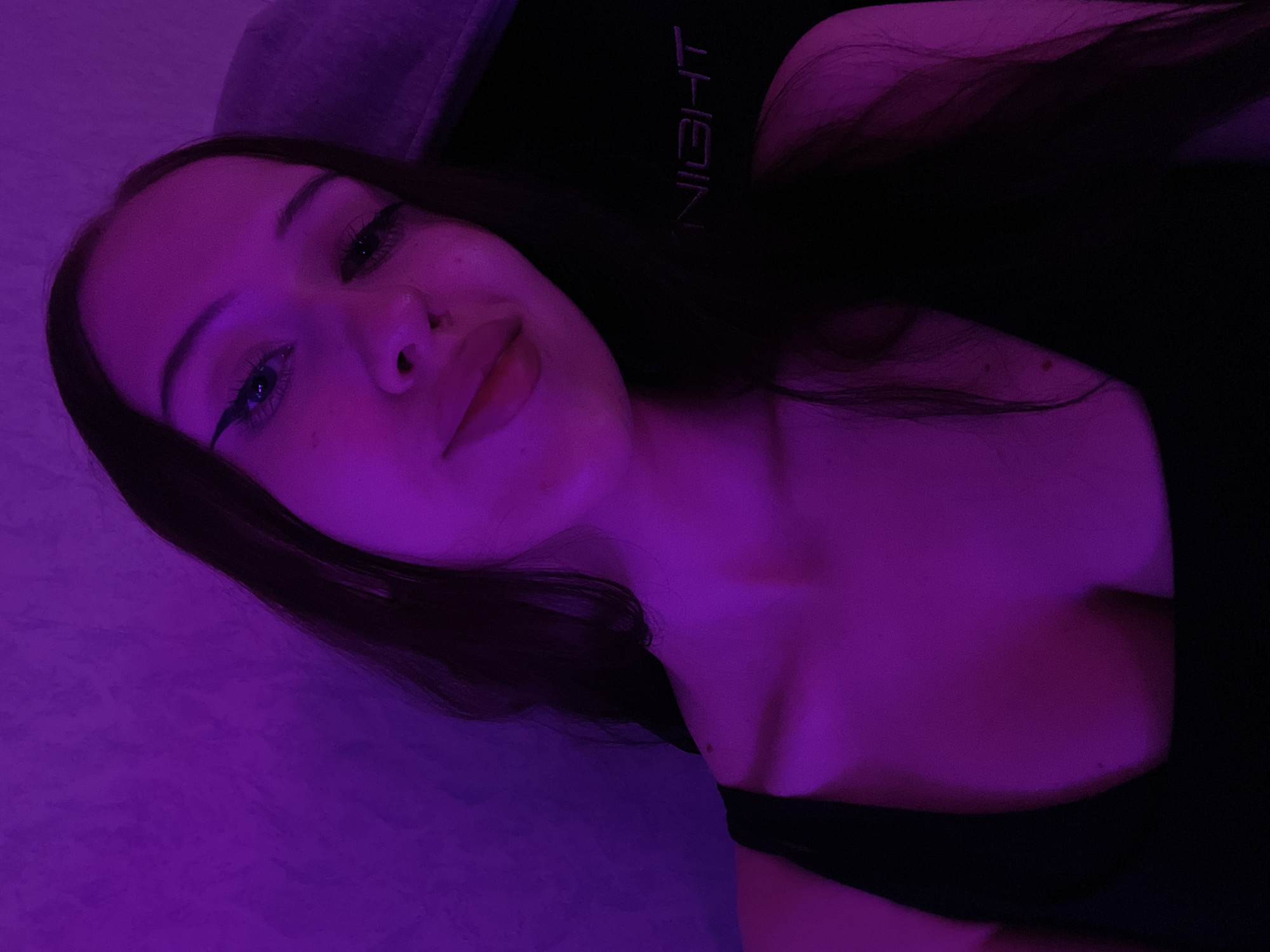 An overhead, close-up, selfie-style shot shows a young woman from the chest up, bathed in purple light. She has long, dark hair that falls over her shoulders and down her back. Her eyes are accentuated with black eyeliner, and her lips are slightly parted. She is wearing a black, low-cut top. Behind her, a textured purple wall is visible, and a portion of a dark chair with the word "NIGHT" embroidered in a lighter thread is also in the frame. The lighting suggests an indoor setting. There is no information to indicate this scene is located in Votkinsk, Russia.