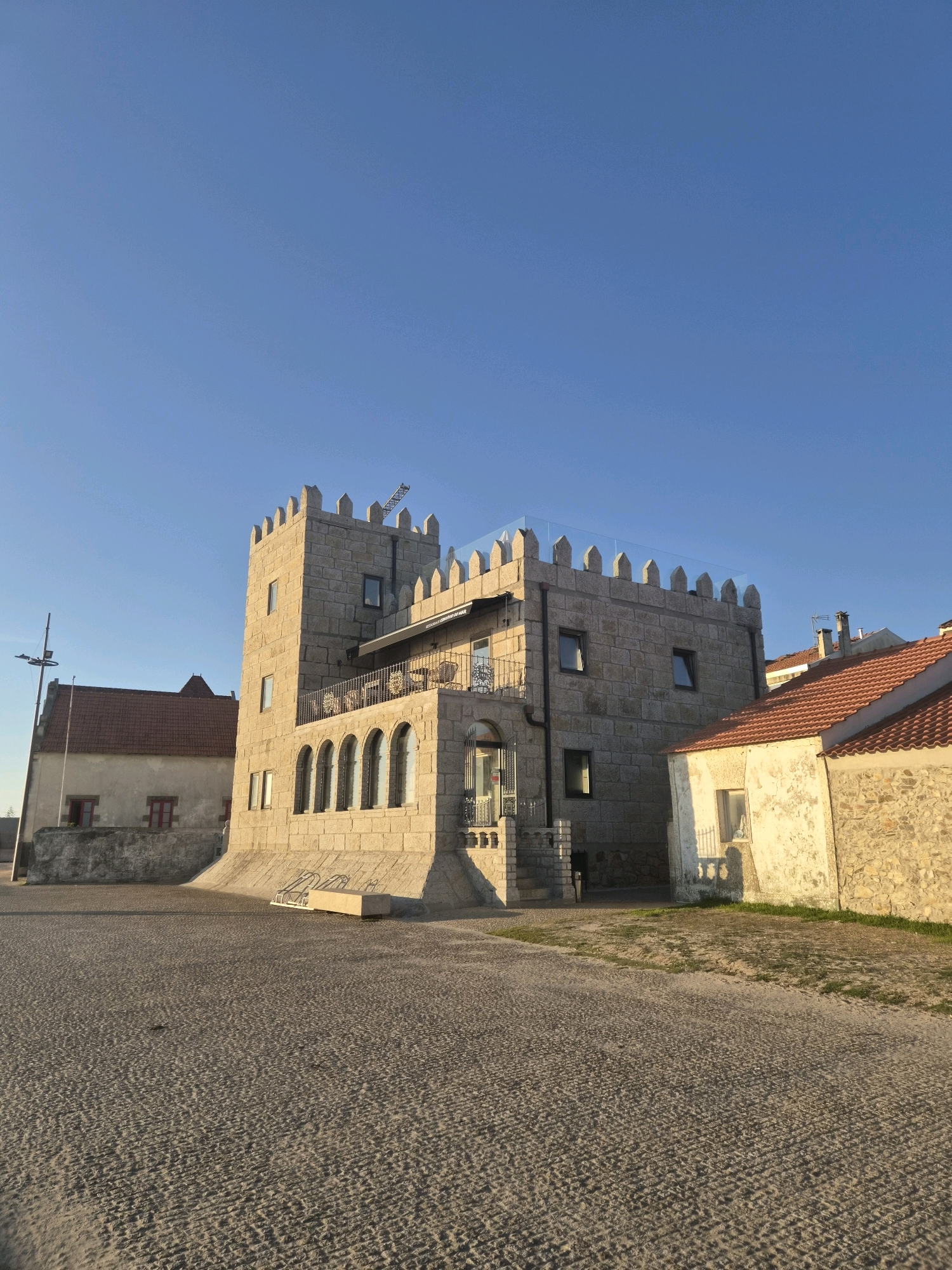 The image shows a view of the Torre de São Lourenço in Esposende, Portugal, a fortified tower built in the 16th century. The tower, constructed of rough-hewn stone blocks, is the central focus of the image. It has a square base and a crenellated parapet along the top. A balcony with wrought-iron railings extends from the main body of the tower. Several arched windows are visible on the lower portion of the tower, and rectangular windows are present on the upper floors. To the left of the tower is a lighter-colored building with a red-tiled roof and red-framed windows. To the right of the tower, another building with a red-tiled roof and a weathered, light-colored facade is visible. The ground in front of the buildings is paved with cobblestones and some patches of sand. The sky above is clear and blue, indicating a sunny day. The lighting suggests it is either early morning or late afternoon, casting long shadows. There are no people visible in the image.