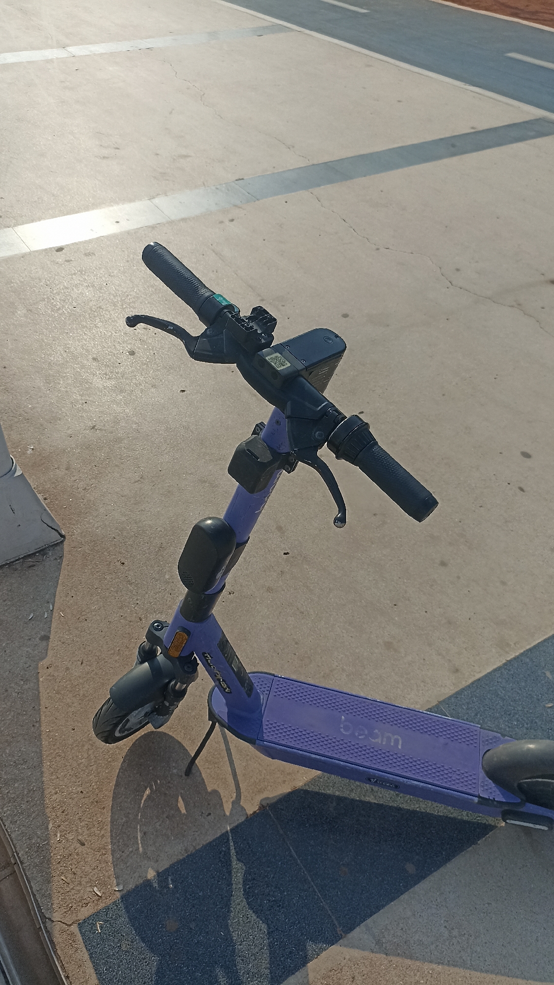 A purple electric scooter is parked on a light-colored paved surface with distinct linear patterns. The scooter is positioned at an angle, with its handlebars prominently displayed in the upper portion of the image. The handlebars are black, featuring textured grips and brake levers. A black digital display is mounted on the handlebars, and a small black box is attached to the vertical stem of the scooter. The deck of the scooter, also purple, has the word "beam" embossed in white. The scooter's front wheel and fender are visible on the lower left, and its kickstand is deployed. Shadows cast by the scooter and an unseen object are visible on the ground. The background consists of a continuation of the paved surface, with varying shades suggesting different materials or textures, and a hint of a reddish-brown area to the far right. The scene appears to be outdoors, under daylight conditions. The image was taken in Yenişehir, Turkey.