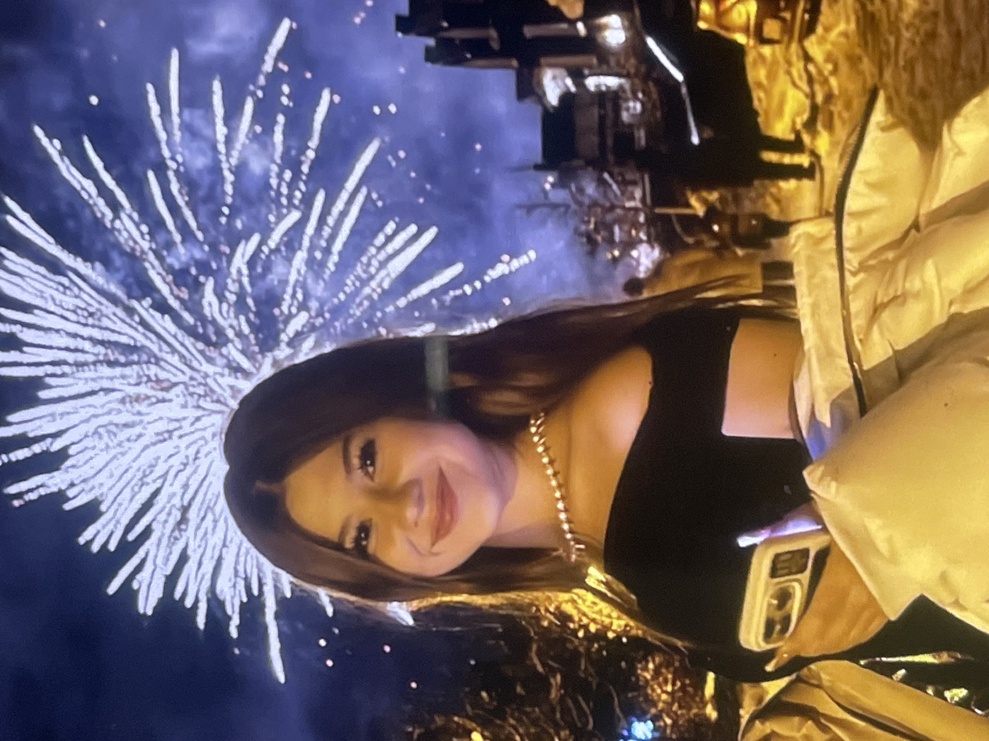 The image, taken in Poltava, Ukraine, captures a young woman smiling at the camera against a vibrant nighttime backdrop of fireworks. The woman, with long, dark hair, occupies the right side of the frame. She wears a dark, possibly black, off-the-shoulder top and a prominent gold-colored chain necklace. In her hand, she holds a white smartphone with a visible multi-lens camera on the back.
Behind her, brilliant white fireworks burst across the dark blue night sky, creating a striking visual effect. To her right, a person wearing a light-colored, possibly cream or white, puffy jacket is partially visible. The far background features blurred, dark structures, likely buildings or trees, illuminated by warm artificial lights, suggesting an urban environment.
The overall scene depicts a celebratory event, likely outdoors at night, given the fireworks display. The woman's smile conveys a sense of happiness and enjoyment, posing for the camera during this festive occasion. No discernible text is clearly visible within the image, apart from faint unreadable characters on the phone screen.