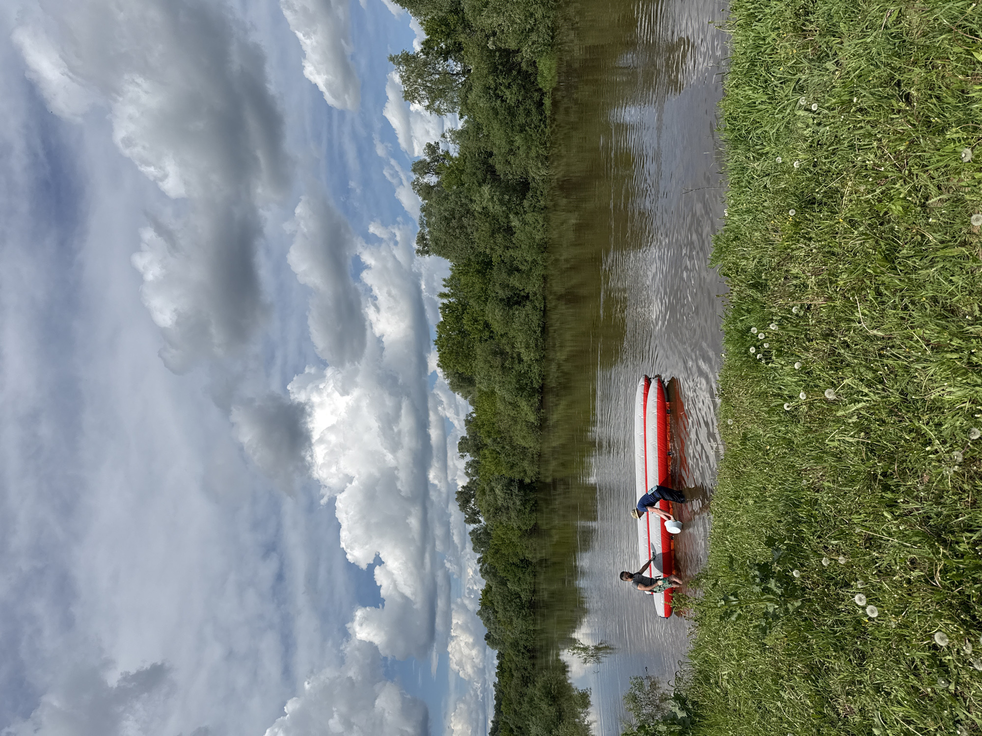 Two people are standing on an inflatable red and white paddleboard in a river, next to a grassy riverbank. Trees line the opposite bank of the river. The sky above is partly cloudy.
While the location requested is Nazyar, Russia, there is nothing in the image to confirm it is that location. It is possible this is not the correct location.