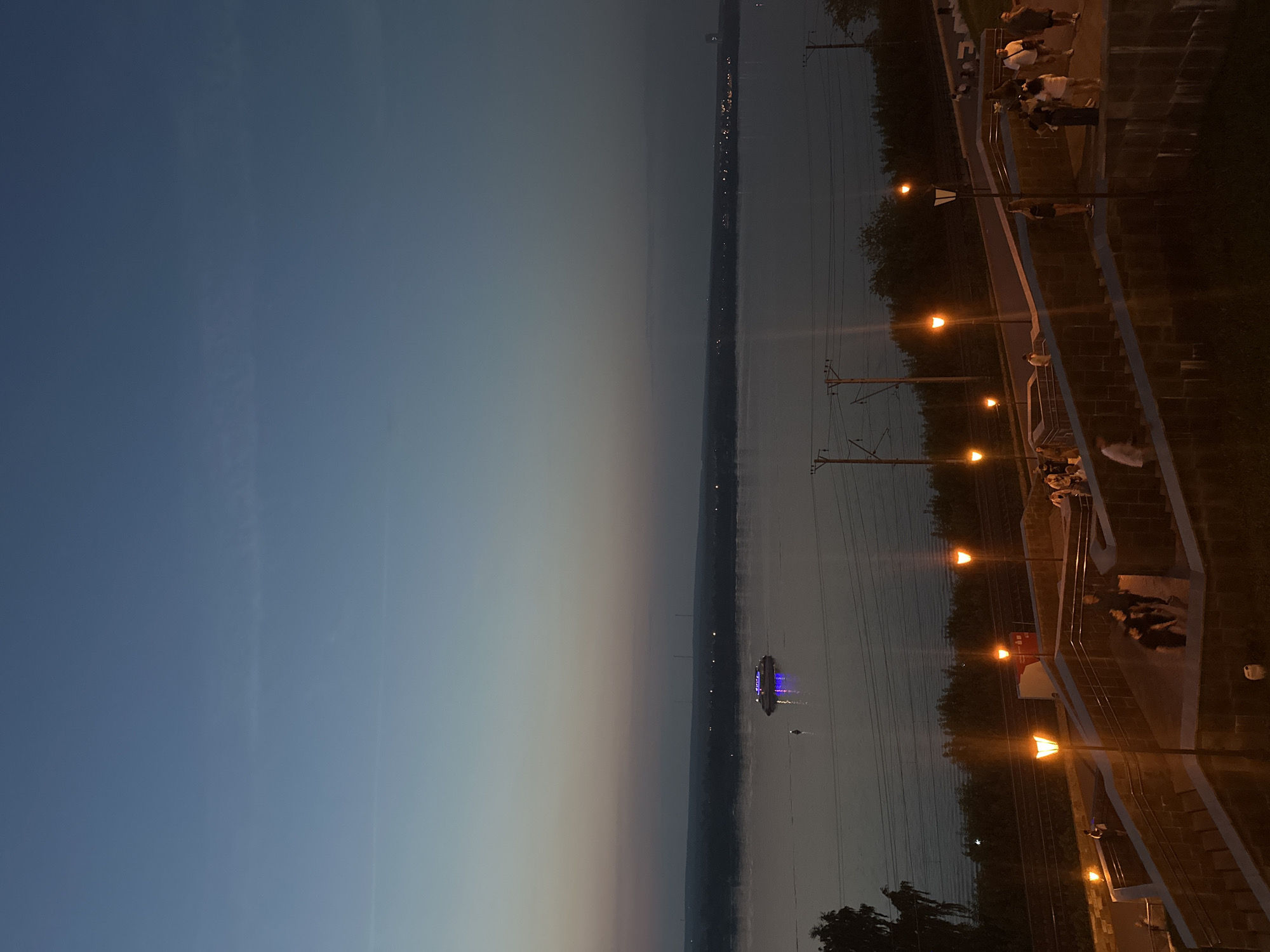 The image captures a dusky twilight scene overlooking the Kama River in Perm, Russia. A wide river dominates the middle ground, with a vessel emitting blue lights visible in the distance. The far bank of the river is lined with a dark silhouette of trees and distant city lights. In the foreground, a set of tiered stairs and pathways are illuminated by warm-toned streetlights, with several people walking and standing on them. Electrical wires and poles are visible between the foreground and the river. The sky transitions from a deep blue at the top to lighter shades of blue and pink near the horizon.