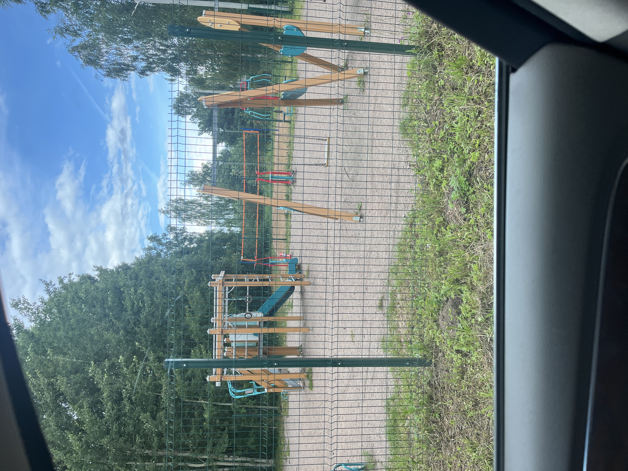 The image shows an outdoor playground enclosed by a green metal mesh fence. The playground surface is covered in light-colored gravel. To the left, there is a wooden and metal play structure with a blue slide and monkey bars. In the center, a volleyball net is visible, suggesting a court. To the right, a swing set with two swings is present, one with a yellow seat and the other with a blue seat. The background is dominated by lush green trees and a bright blue sky with scattered white clouds. The image appears to have been taken from inside a vehicle, with the dark interior of a car visible in the foreground. The scene is a typical public park or recreational area. This location is identified as being in Vladimirovka, Russia.