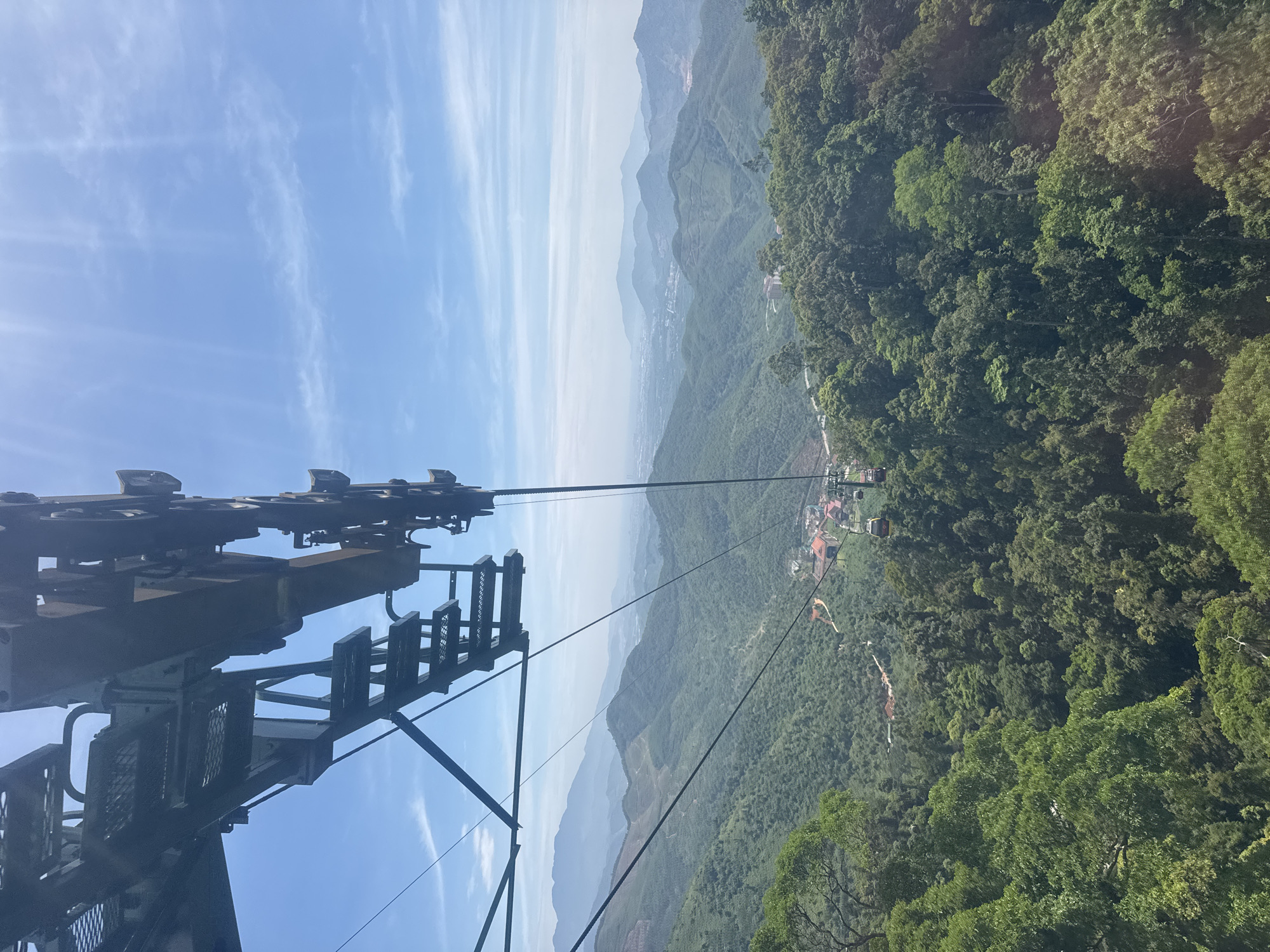 The image depicts a view from a cable car ascending a mountain in Đà Nẵng, Vietnam. The foreground features the metal structure of the cable car system. The midground shows a dense, green forest covering the mountainside. A smaller village or resort area is visible amidst the trees. In the background, a hazy mountain range fades into the distance under a blue sky streaked with light clouds.
