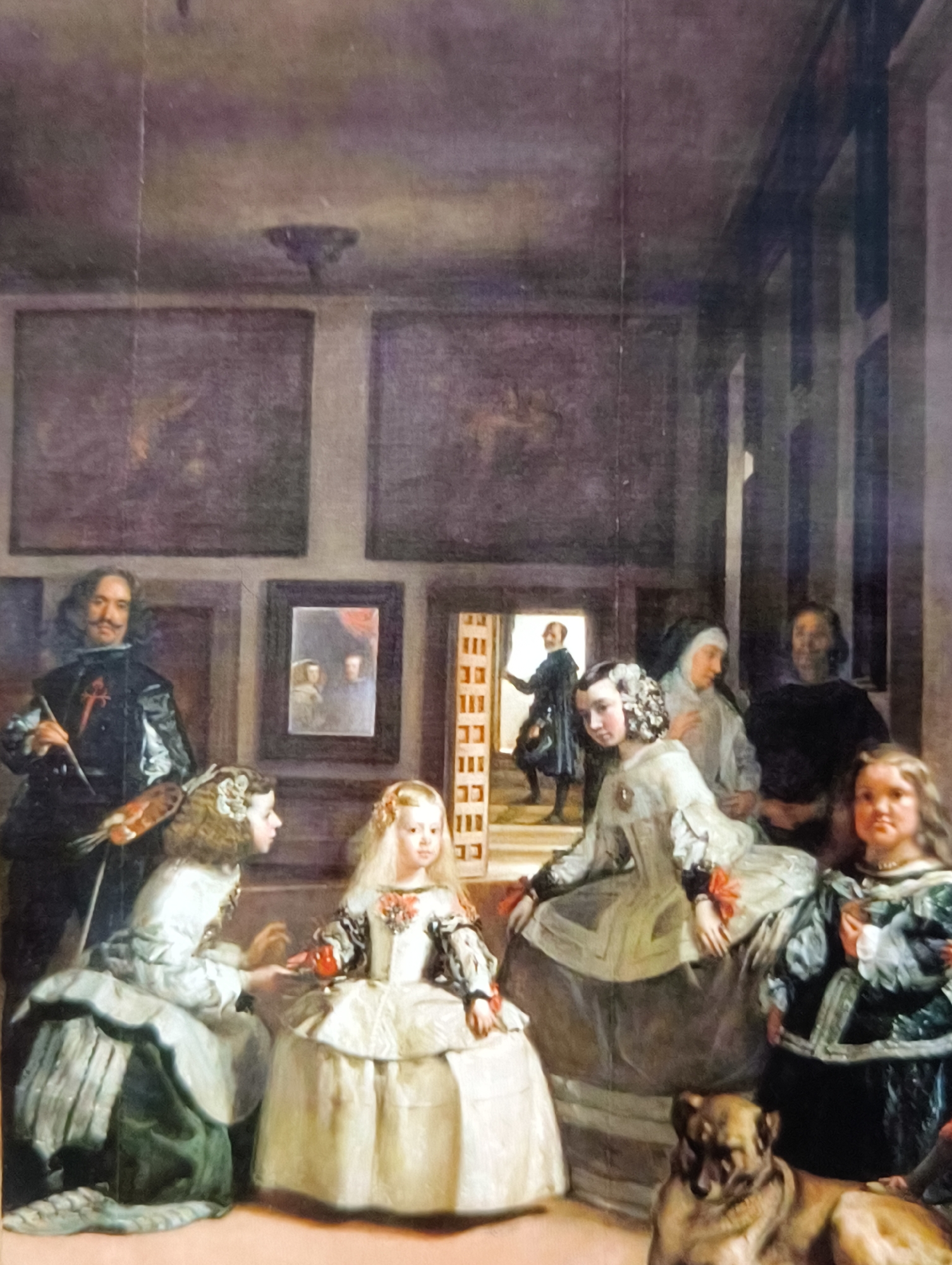 The image depicts a scene within a dimly lit interior, likely a palace chamber. The focus is on a group of individuals, primarily women and children, in elaborate 17th-century attire.
In the foreground, a large dog with tan fur is lying down, looking towards the viewer. To the left, a man with a mustache, dressed in a black suit with a red sash, stands holding a paintbrush and a palette. He appears to be the artist.
In the center, two young girls are prominent. The girl on the left, facing away from the viewer, wears a white dress and has ornate hair. The girl on the right, facing the viewer, also wears a white dress with intricate details and a pearl necklace.
Behind them, several other figures are visible. A woman in a nun's habit stands to the right of the central figures. To her right, a dark-haired woman with a stern expression is partially visible. Further back, a man in dark attire is seen through an open doorway, walking away from the room.
The background features large paintings hanging on the walls, suggesting an art-filled environment. The room appears spacious, with tall columns and archways visible in the deeper recesses. The overall lighting is subdued, with light sources not directly apparent but contributing to the chiaroscuro effect. The scene is densely populated with figures and objects, creating a sense of depth and activity within the regal setting.