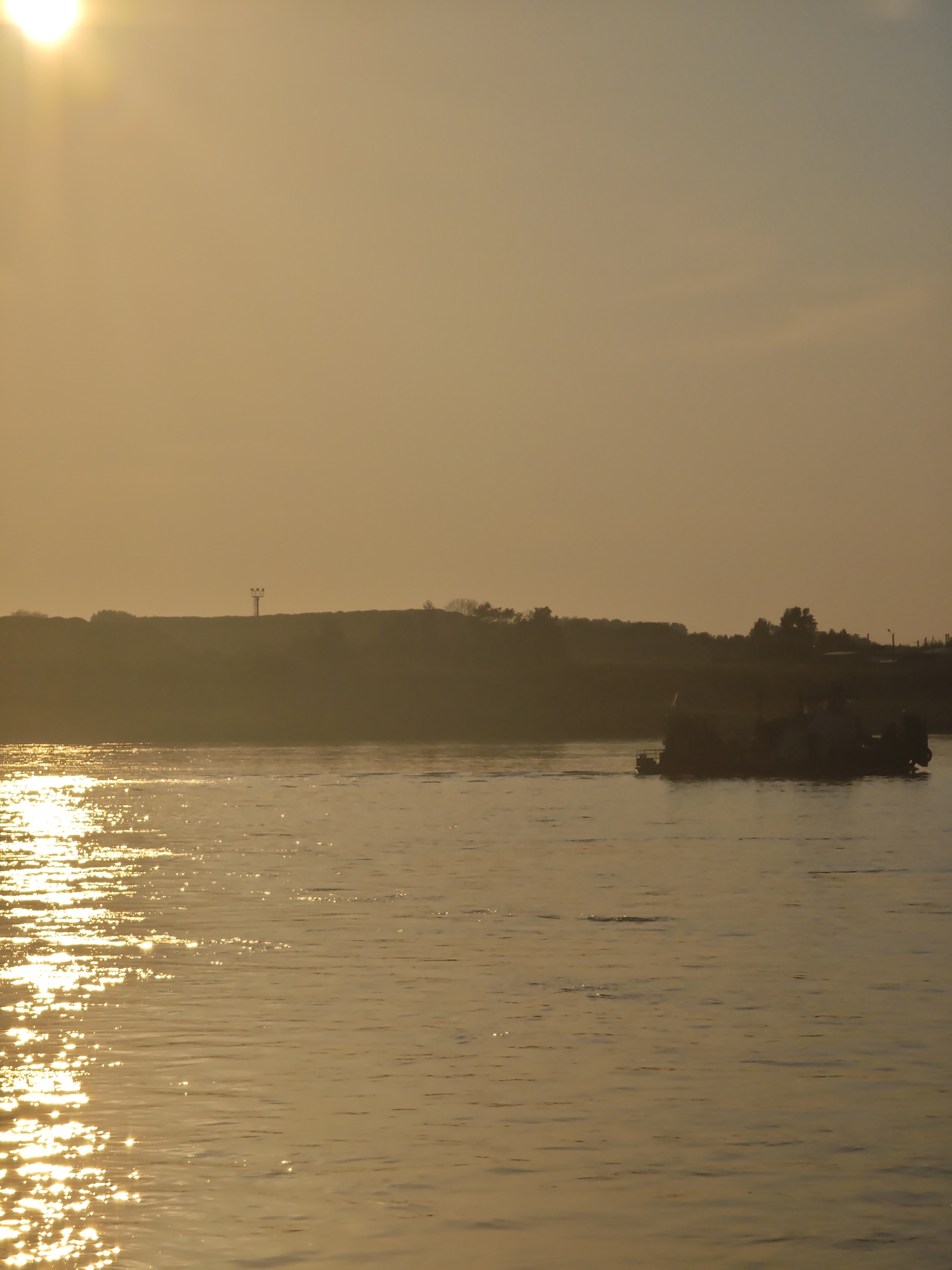 A wide shot captures a body of water bathed in the warm, golden light of a setting sun. The sun, partially visible in the upper left corner, casts shimmering reflections across the water's surface, creating a pattern of light and shadow. In the distance, a silhouetted shoreline rises, dotted with trees and a single tower-like structure. A barge, also silhouetted, navigates the water from right to left. The sky is a pale orange, with soft clouds present. The scene appears to be a natural landscape. It is not possible to determine the specific location within Lesosibirsk, Russia, from the visual information provided.
